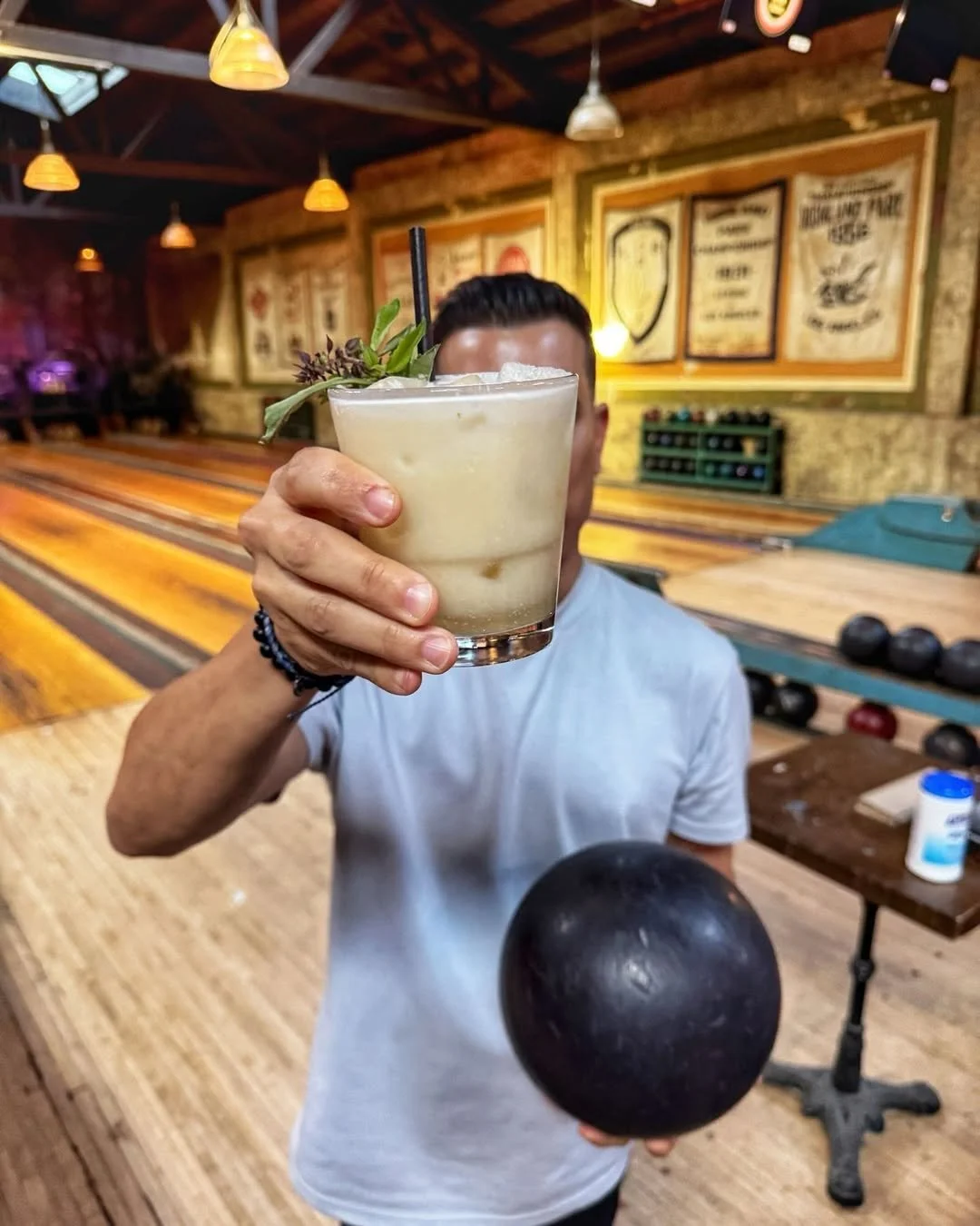 A guest holds a craft cocktail in one hand and a bowling ball in the other inside Highland Park Bowl, capturing the venue's social bowling experience.