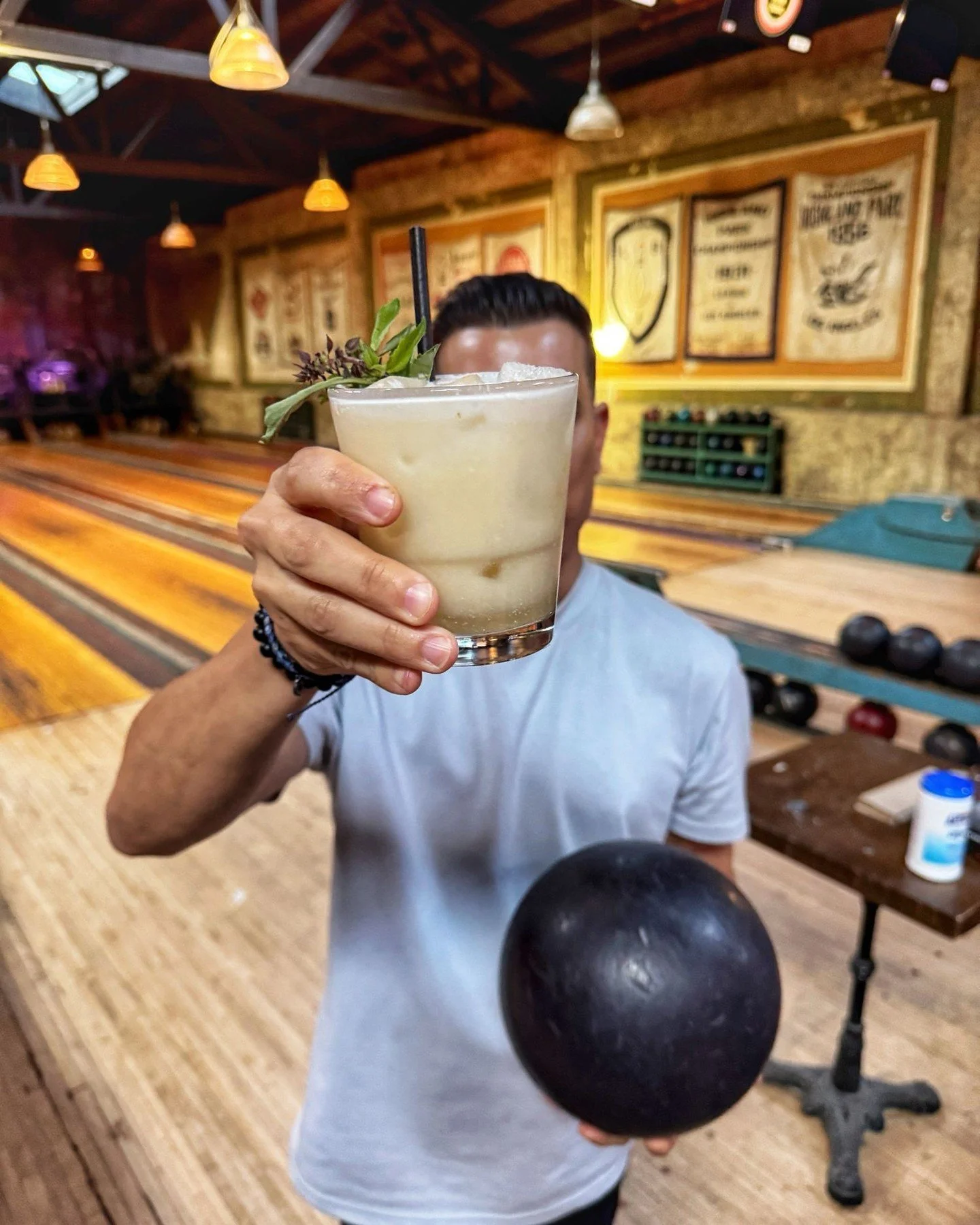 Person holding a cocktail glass and a bowling ball inside a vintage bowling alley at Highland Park Bowl.