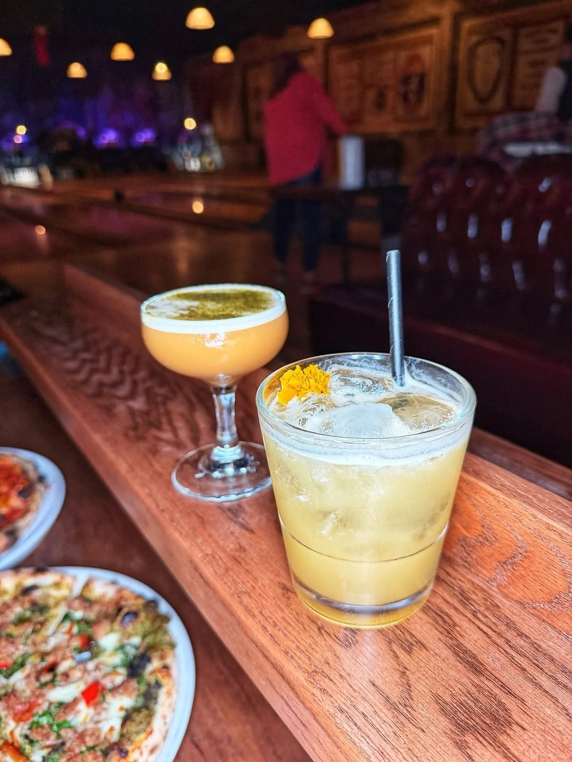 Two craft cocktails resting on a wooden bar with Highland Park Bowl's historic bowling lanes in the background.