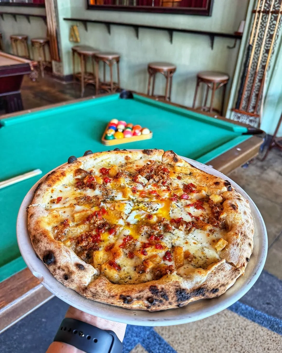 Artisan pizza held in front of a pool table with racked balls at Highland Park Bowl.