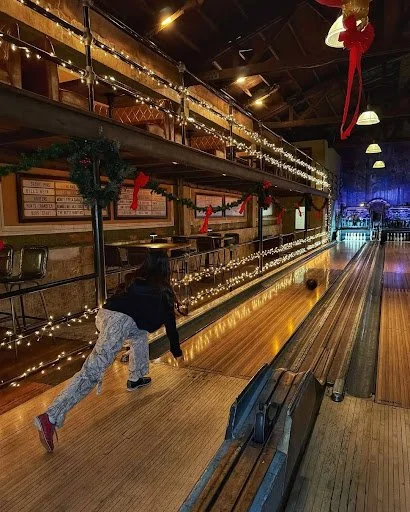 Woman bowling on a warmly lit lane at Highland Park Bowl with string lights and festive décor.