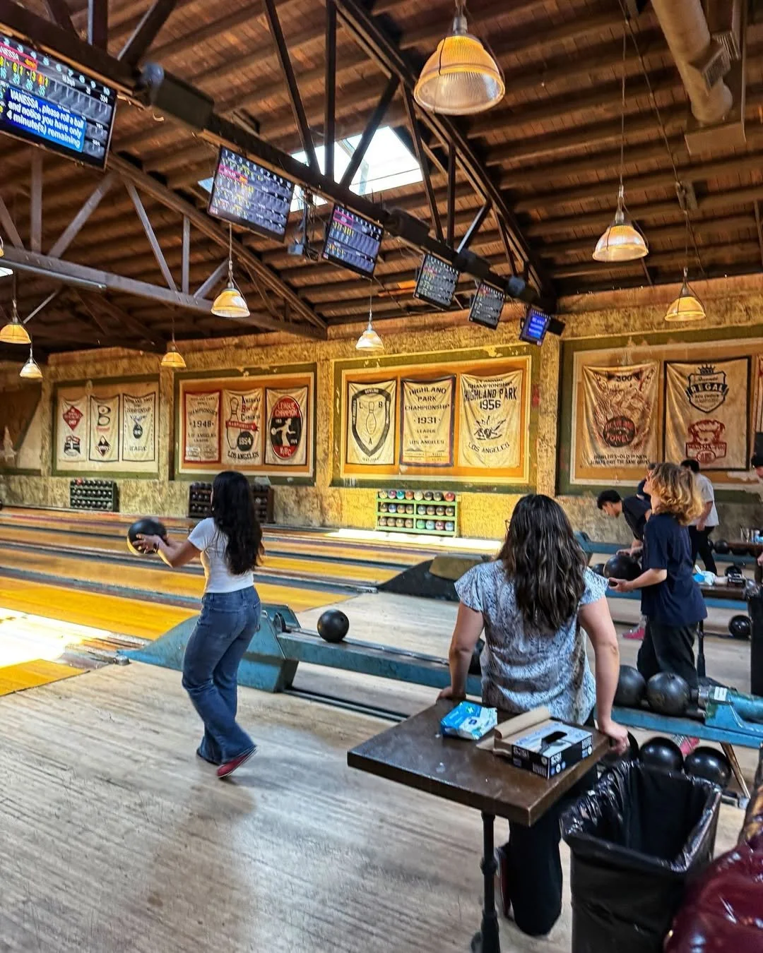 People bowling inside Highland Park Bowl with wooden lanes, hanging lights, and digital score screens.