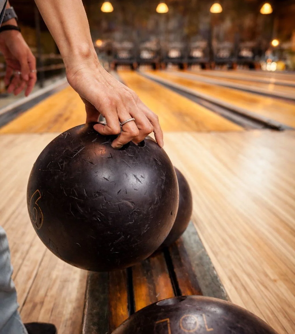 Hand holding a bowling ball at the lane with pins and lights in the background.