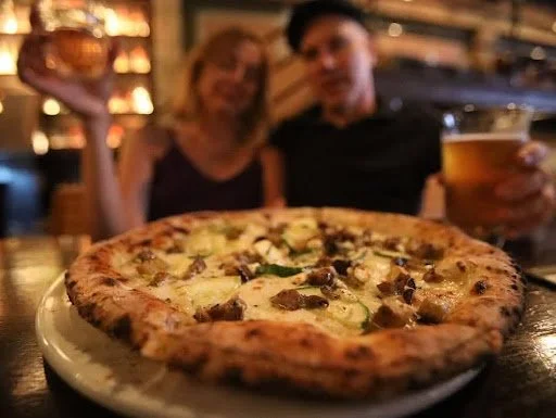 Close-up of pizza in the foreground with a blurred couple holding drinks at Highland Park Bowl.