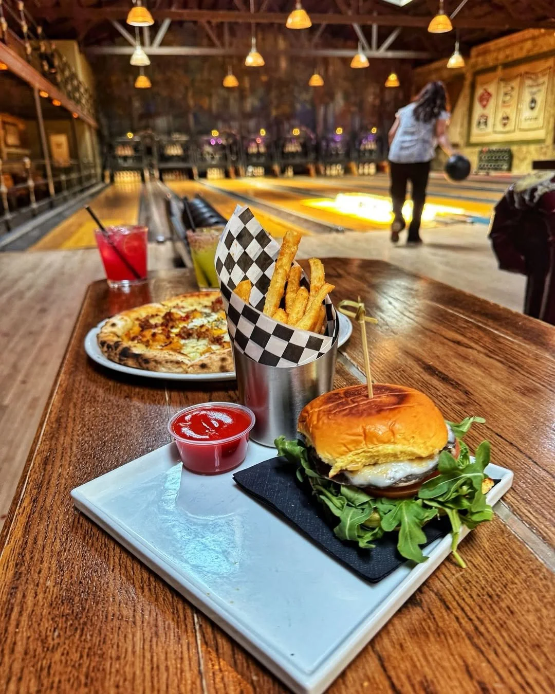 Burger, fries, pizza, and craft cocktails on a wooden table with a bowler in the background at Highland Park Bowl.