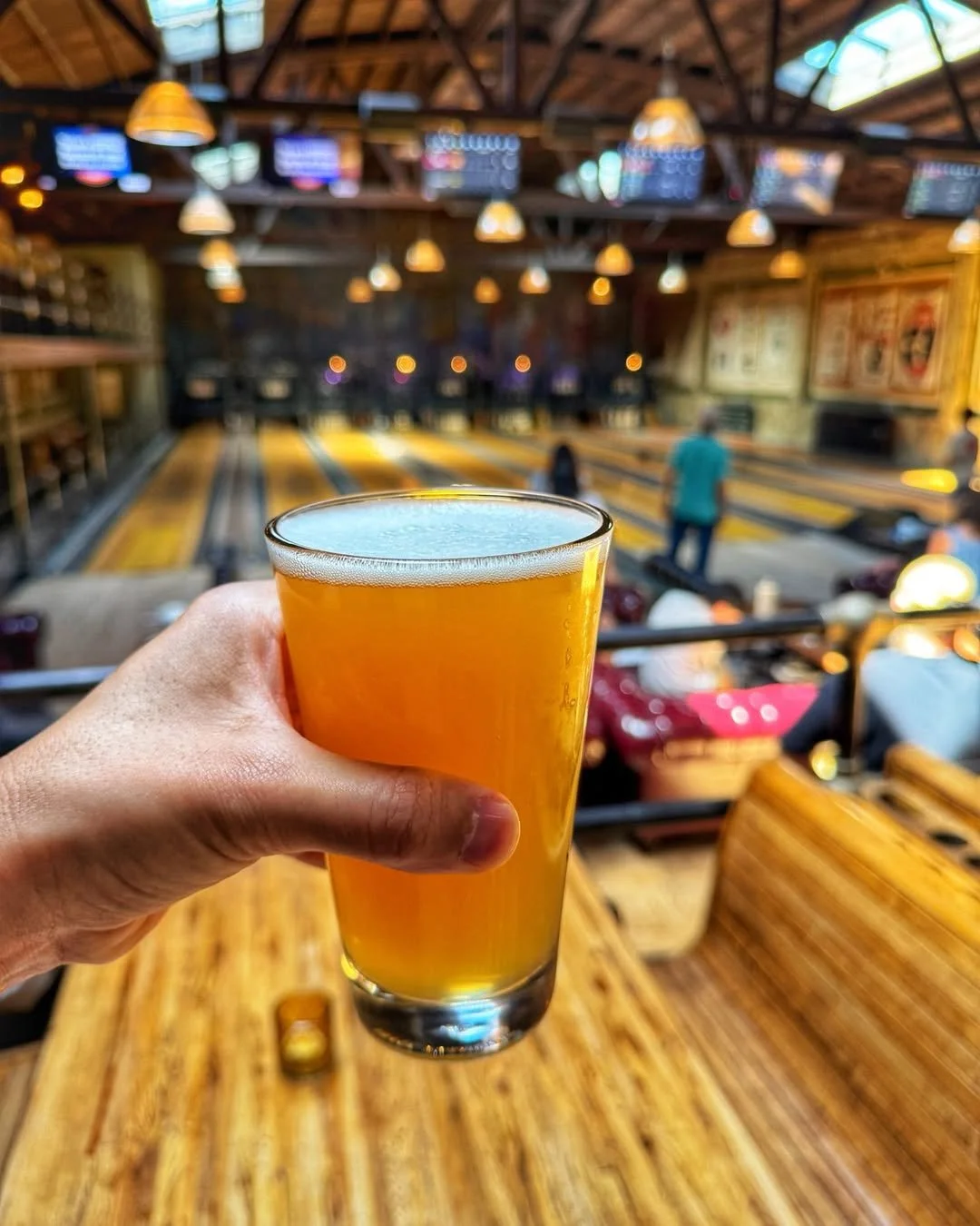 Hand holding a glass of beer with bowling lanes visible in the background at Highland Park Bowl.