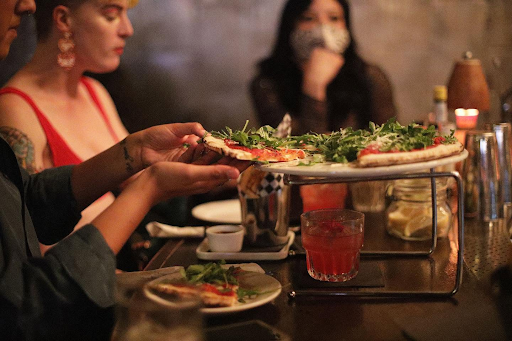 Friends enjoying pizza, cocktails, and conversation at a table inside Highland Park Bowl during a late-night outing.