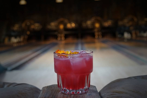 Bright pink craft cocktail garnished with a flower at Highland Park Bowl with bowling lanes in the background.
