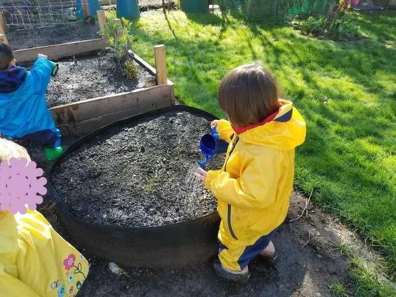 ID: A child in a yellow rain suit uses a small water can to water a garden bed.