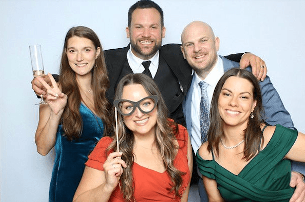 Group of five young adults at a celebration, smiling, with one woman holding a drink and another with costume glasses, dressed in semi-formal attire, in front of a light blue background.