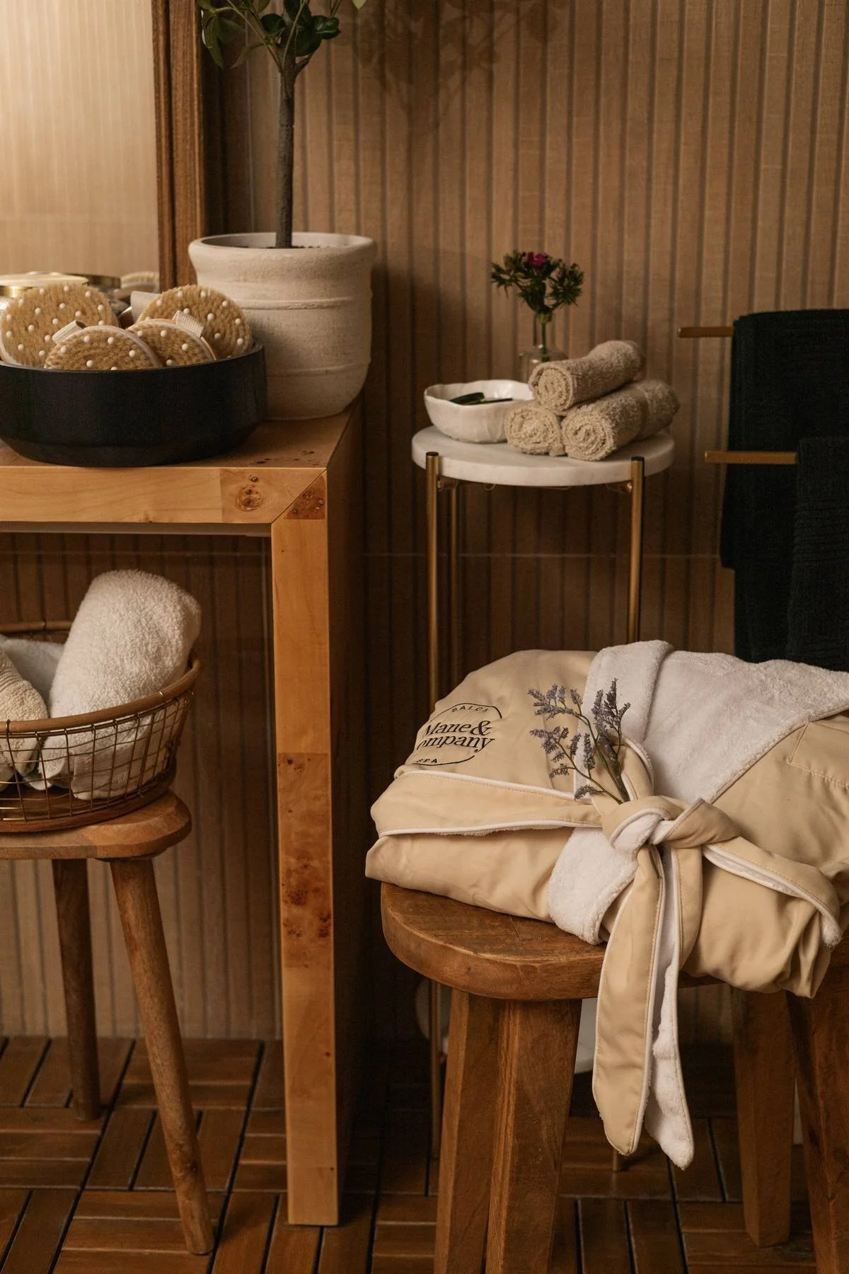 Spa or massage room setup with rolled towels, a tote bag, lavender sprigs, and cleaning cloths on wooden and marble furniture against wood-paneled walls.