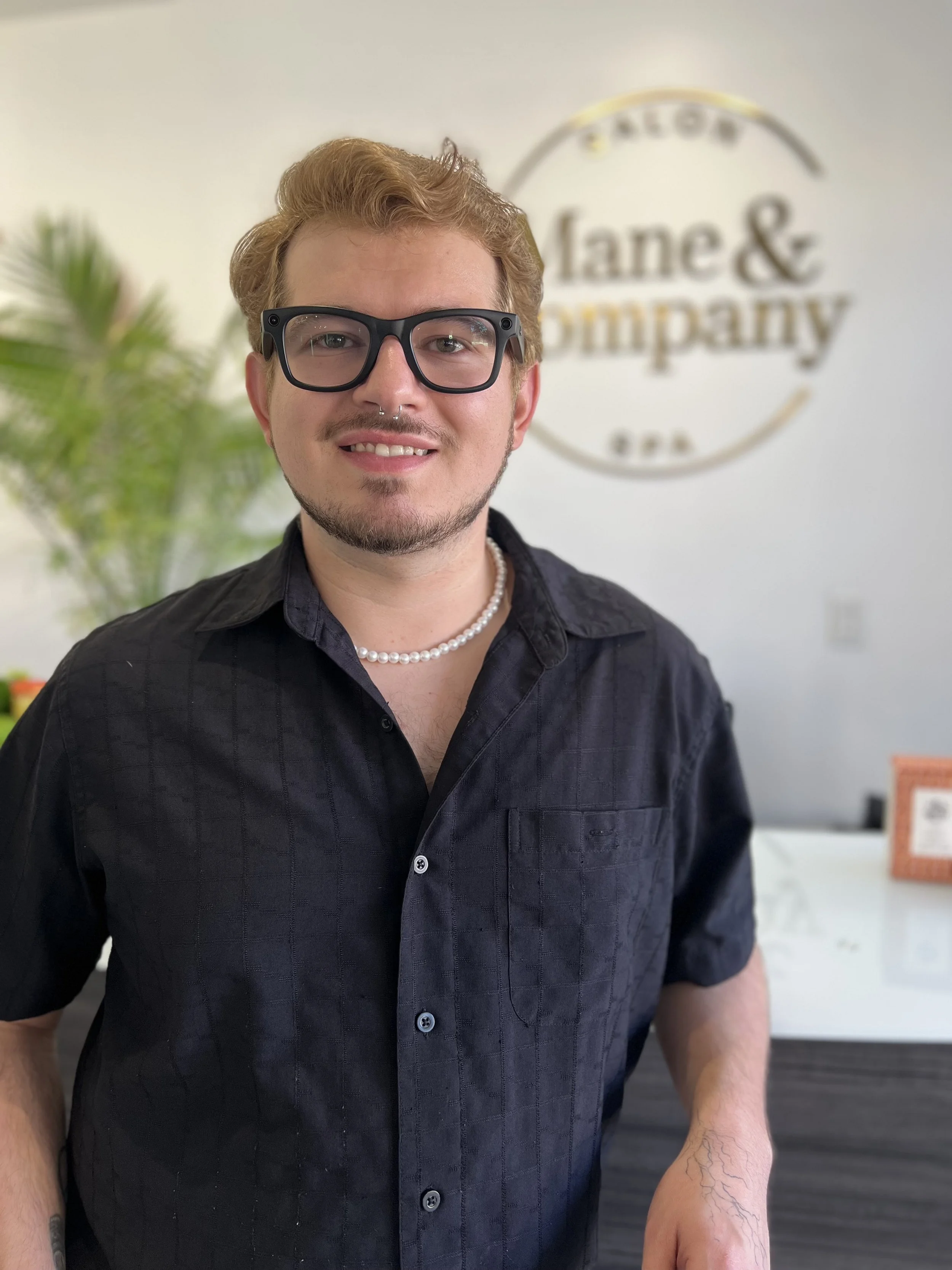 A young man with blonde hair, beard, and light skin sitting in a black salon chair inside a hair salon, looking at the camera with a slight smile, wearing a black shirt and jewelry.