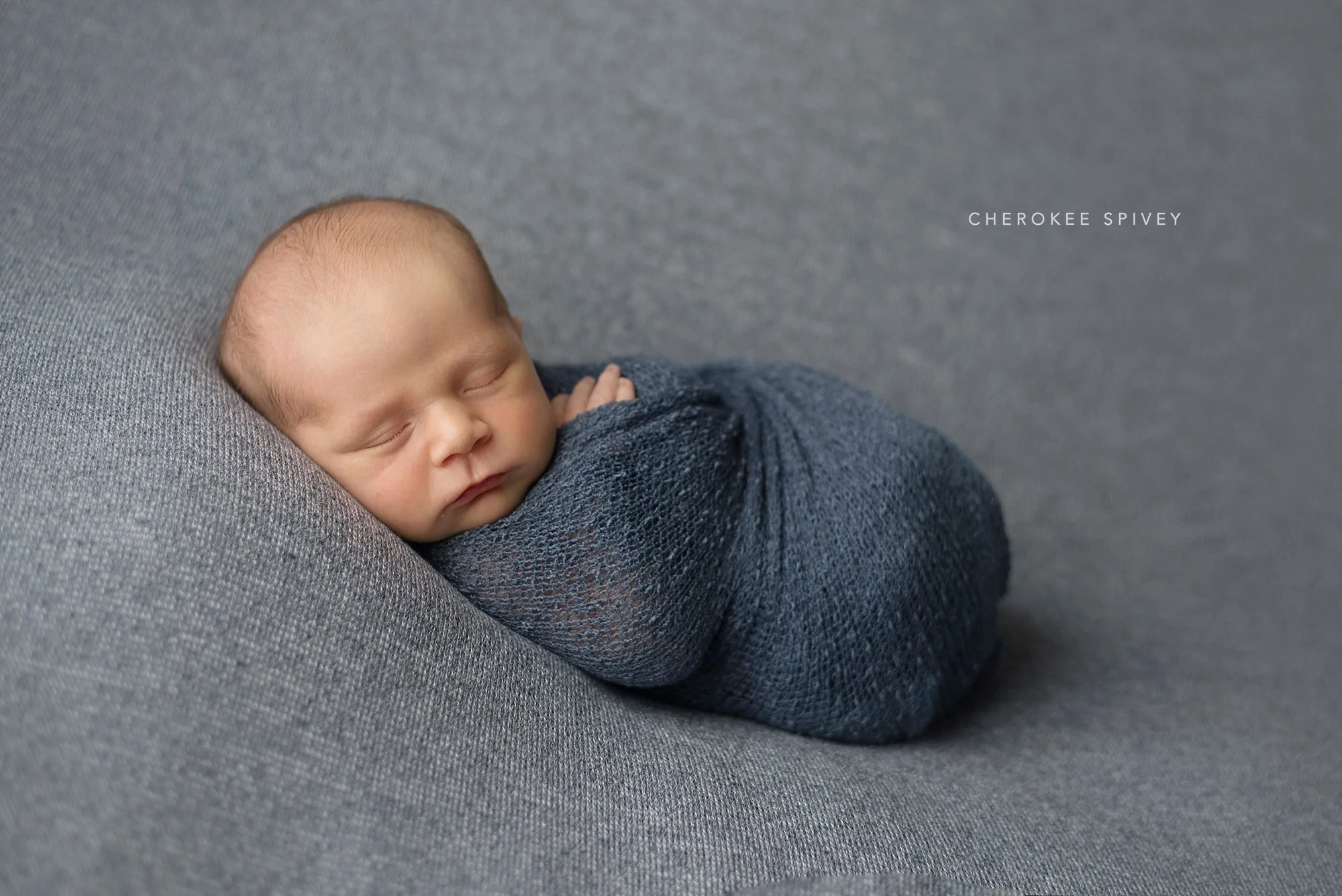 Wee baby boy wrapped in dark gray fabric, reclining on light gray background, his sleeping face so delicate.