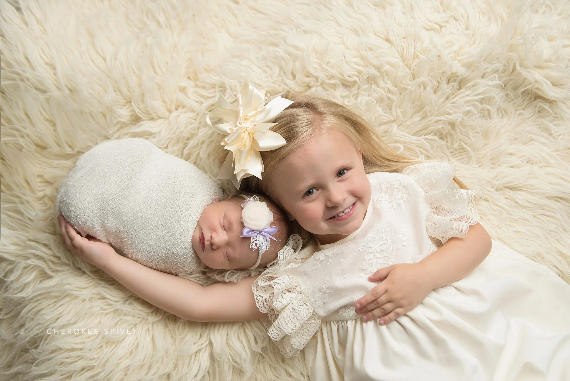Smiling two year old girl laying on an ivory fur, is dressed in a fancy lace ruffled dress, her newborn sister wrapped in white, their faces close together