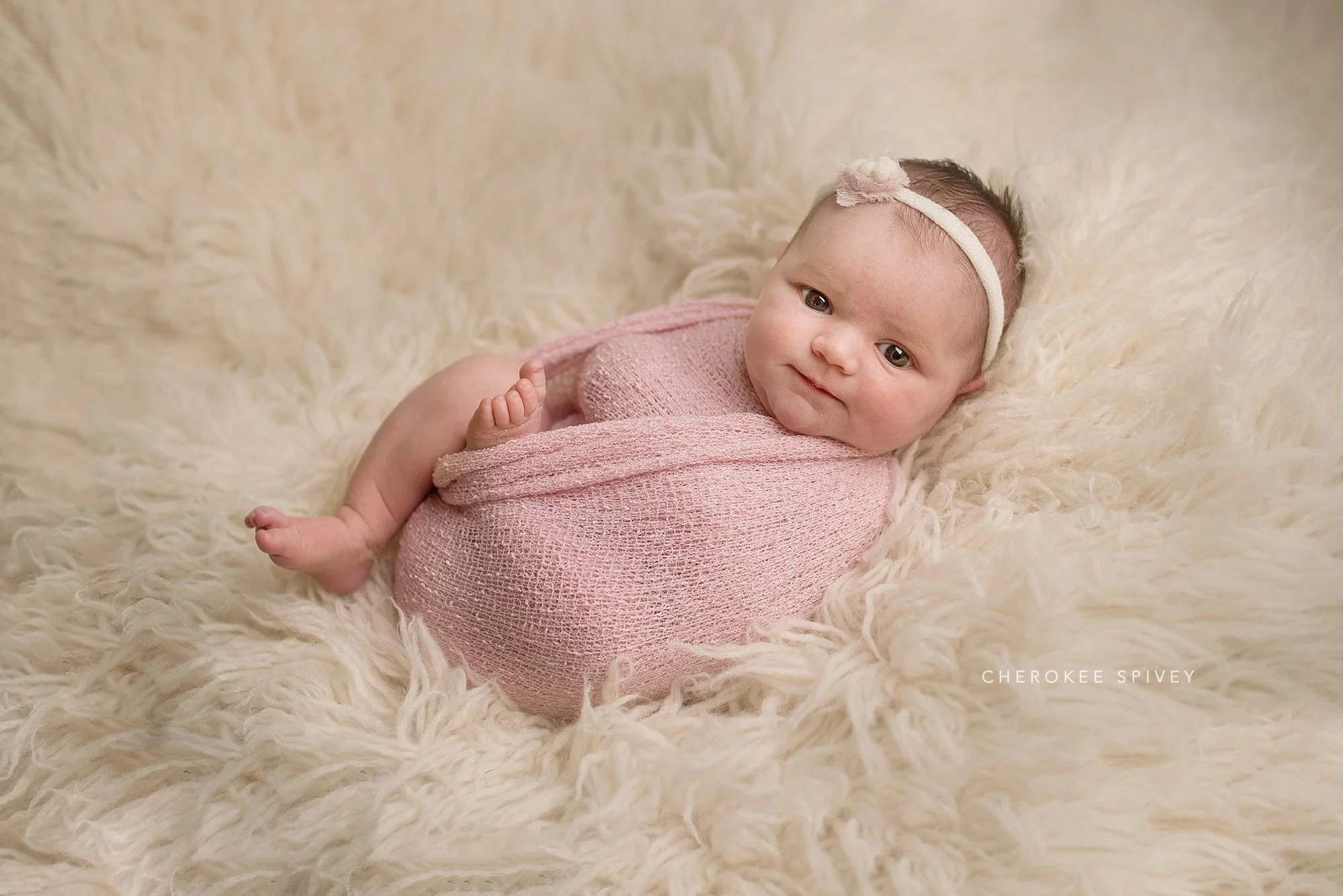 Alert, bright eyed baby girl, tiny feet escaping out of light pink wrap, lying contentedly on her back on an ivory fur.