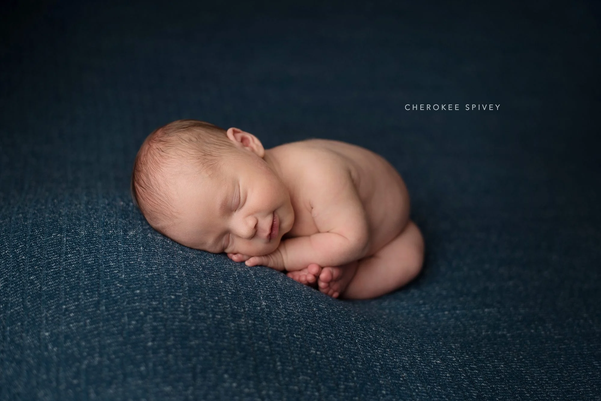 Darling tiny baby boy, curled up naked on his tummy so that his hands are under his cheek, head turned to the side, and feet peeking out. He is sleeping on a navy blanket.