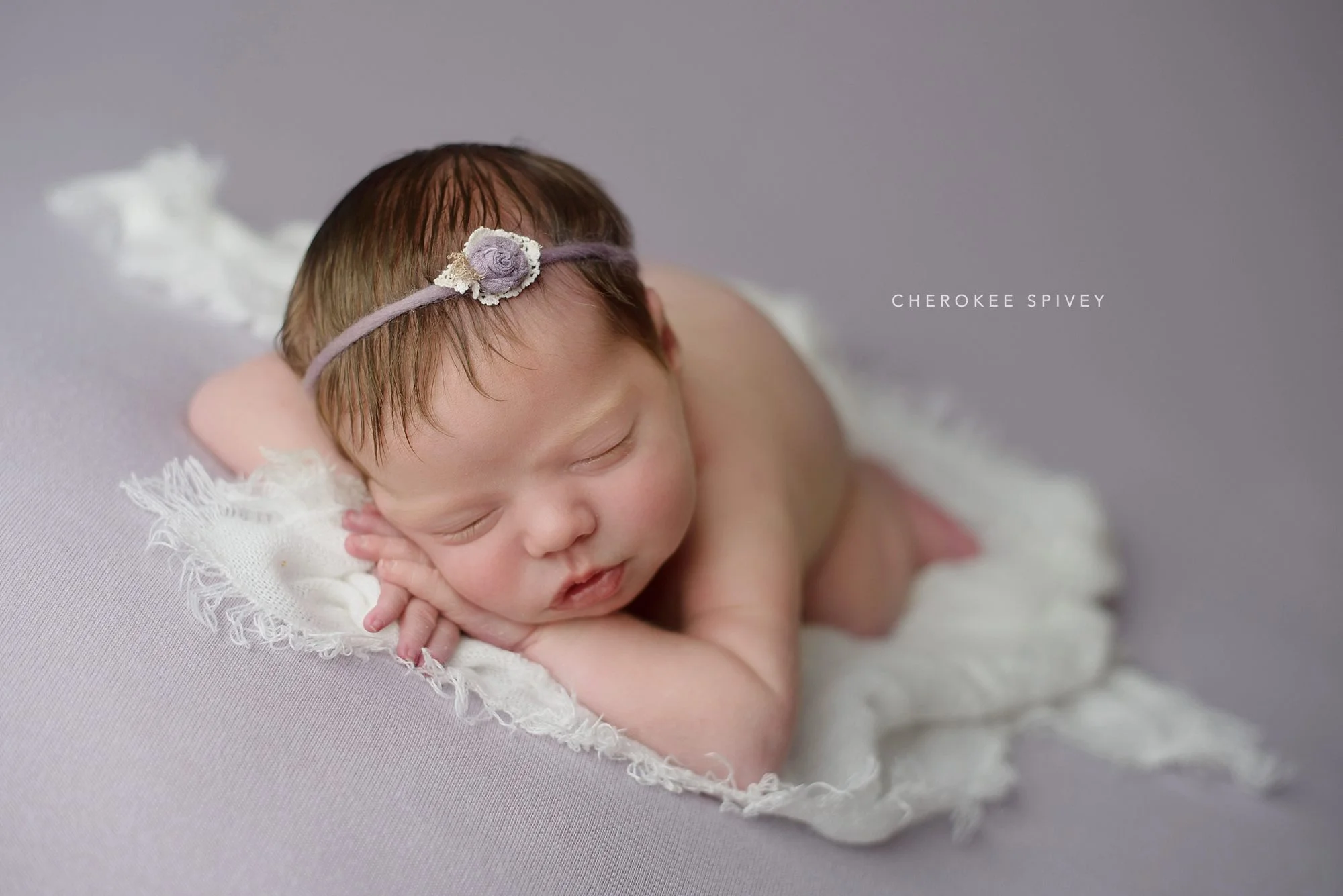 Newborn girl with a lot of dark hair tamed by a flower headband, is laying on her stomach, arms up and her head resting on her hands, on a beautiful fringed scarf.