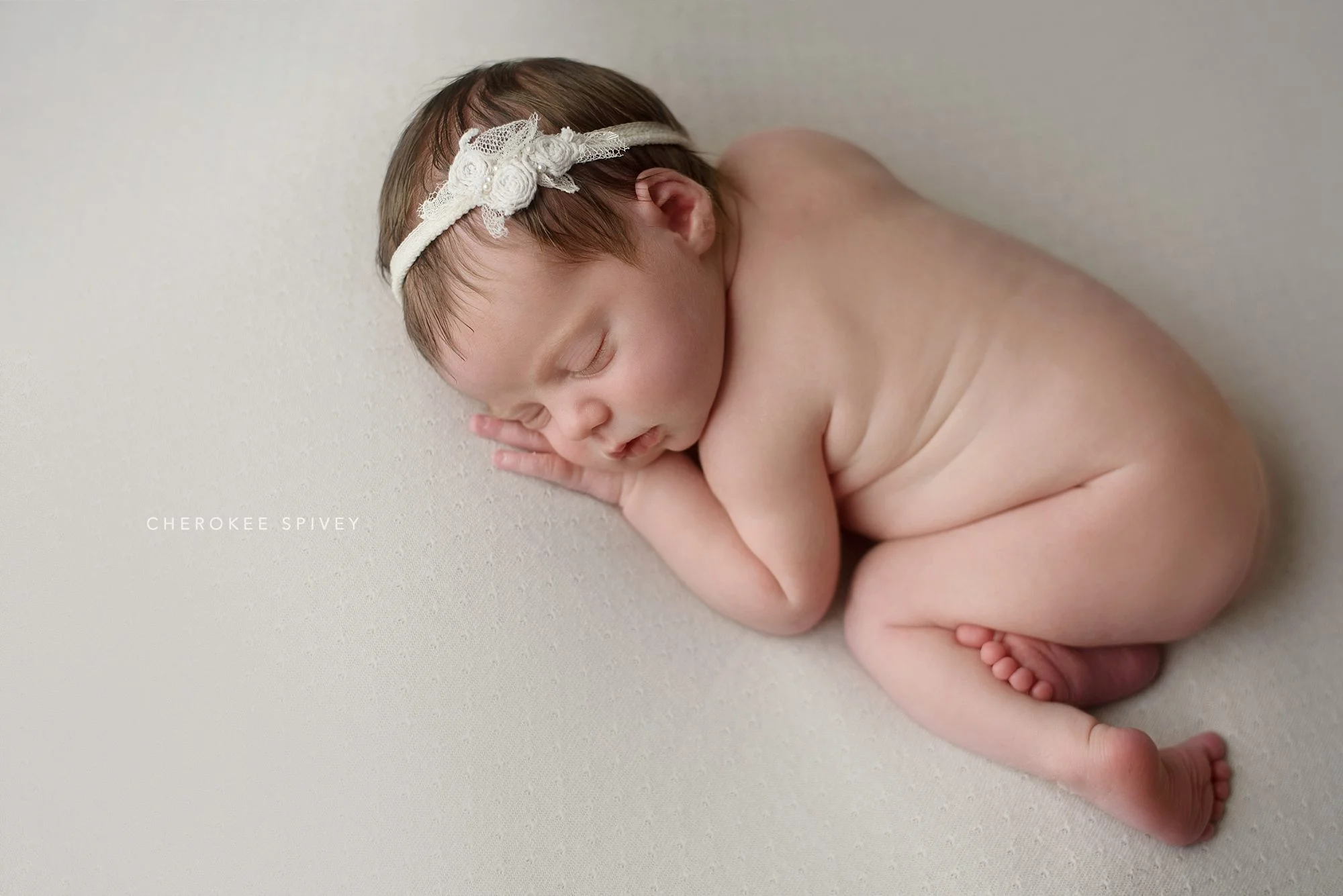 Sweet naked baby girl, white flowers and lace headband accenting her dark hair, is curled up sleeping, her little toes of one foot peeking out from the fold of her other leg.