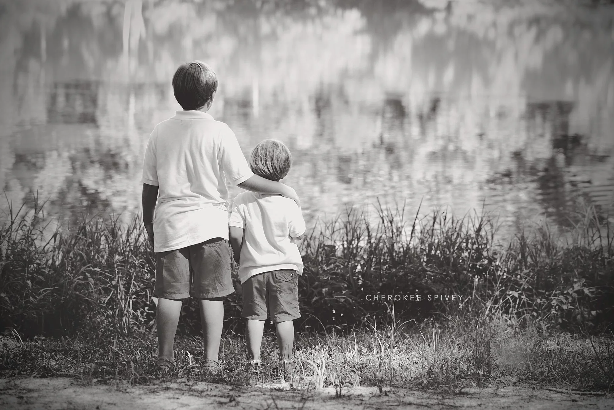 Black and white image of brothers taken from the back, the older boy with his arm protectively around his sibling, at waters edge, as they gaze toward the pond.