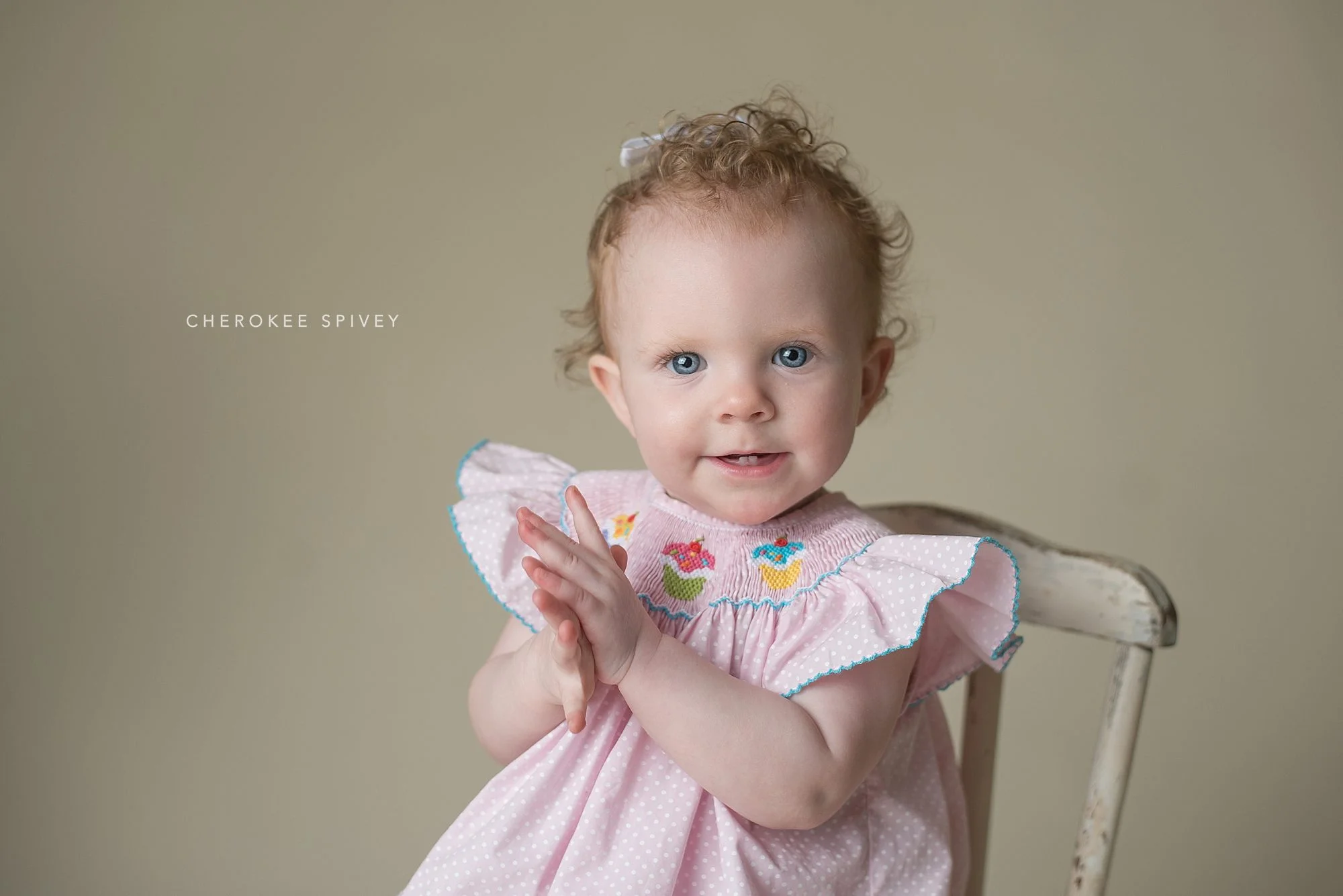 Darling blue-eyed girl sitting in a white chair in a pink dress and delightedly clapping her hands, her smile showing two bottom teeth.