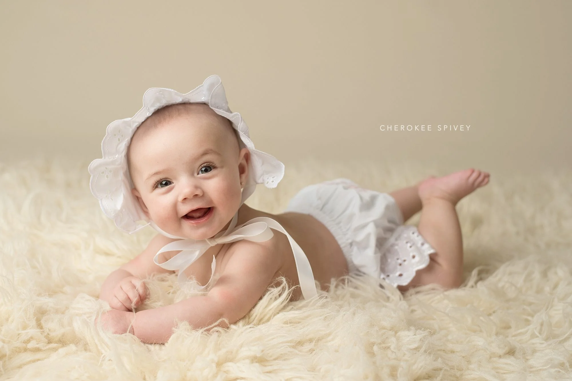 A darling 4 month old girl dressed in a white eyelet bonnet and bloomers, lying on her tummy and pushing up, smiles brightly with her sparkling eyes and toothless grin.