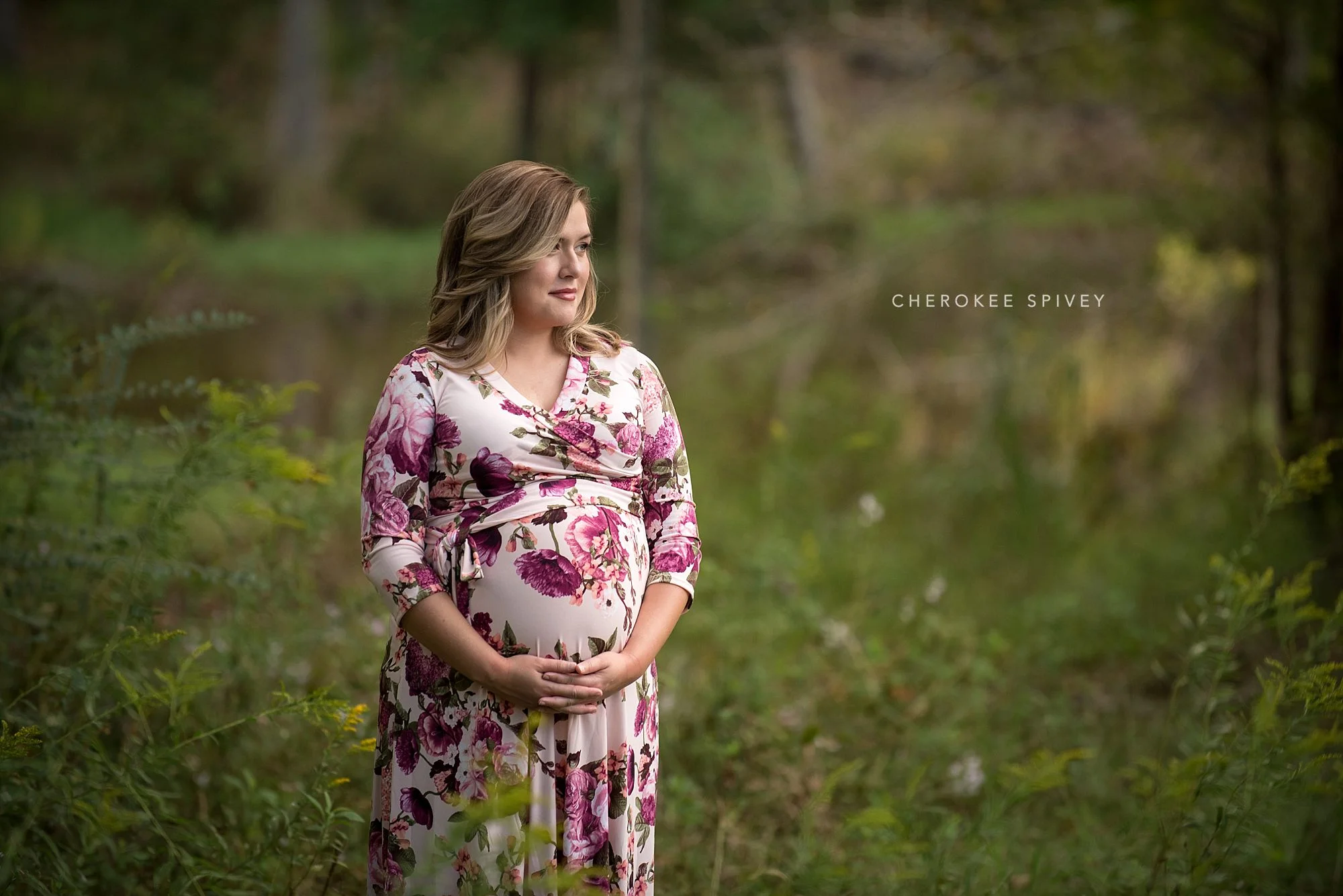 woodland setting with a pregnant woman in a cream and rose floral dress dreamily holding her belly