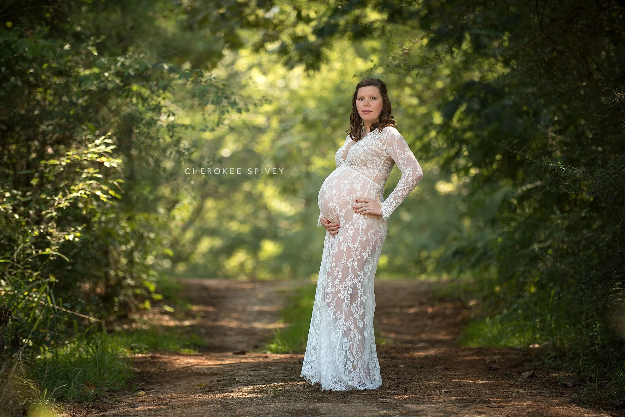 gorgeous woman in a lace gown holding her baby bump surrounded by a tunnel of sunlit trees