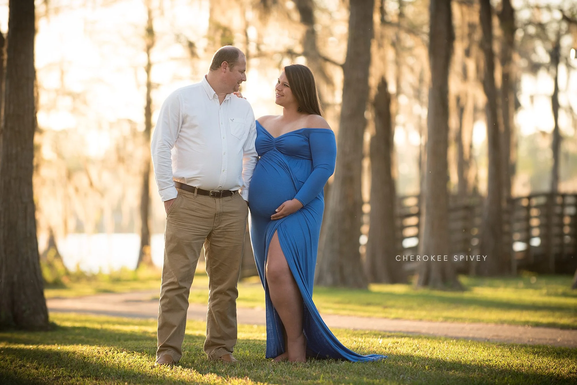 pregnant woman in a striking blue wrap dress gazing into her husbands eyes with a lake and mossy cypress trees in the background