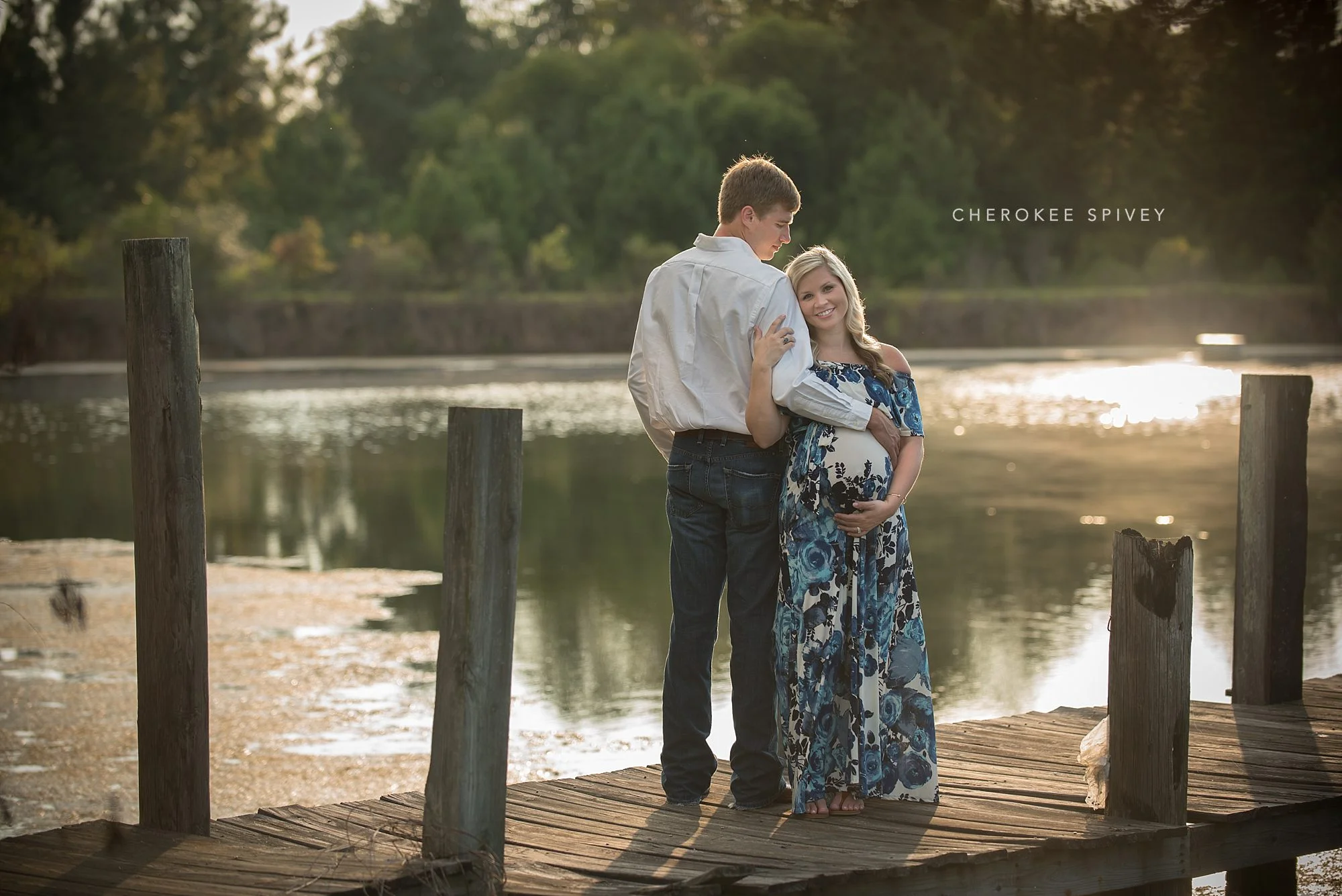 Pregnant woman in a blue maxi-dress, holding her big belly, next to her husband on a pier, with a glistening lake in the background