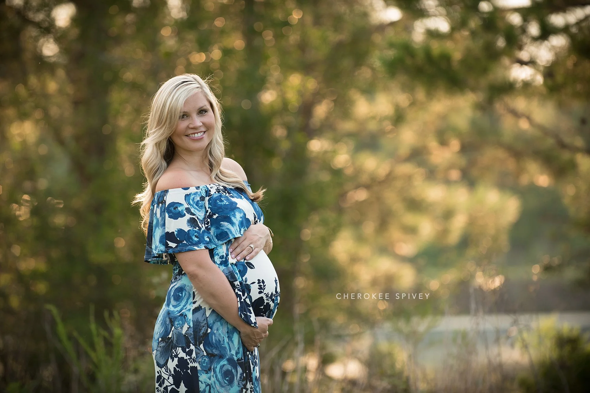 Happy, smiling, young pregnant woman in a blue floral maternity dress in a lovely outdoor setting.