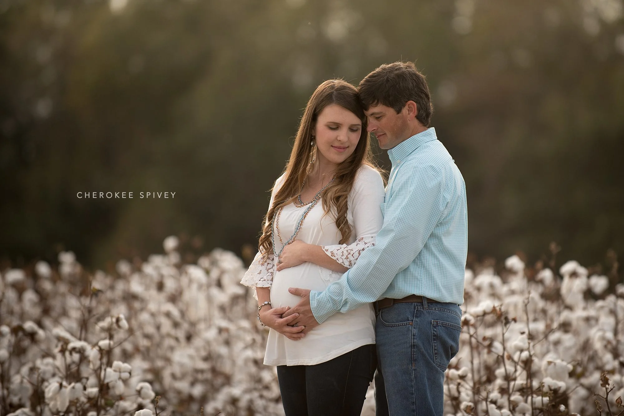 Maternity pictures in a cotton patch! Pregnant brunette young woman in a white shirt affectionately hugged from behind by her good looking husband rubbing her big belly.