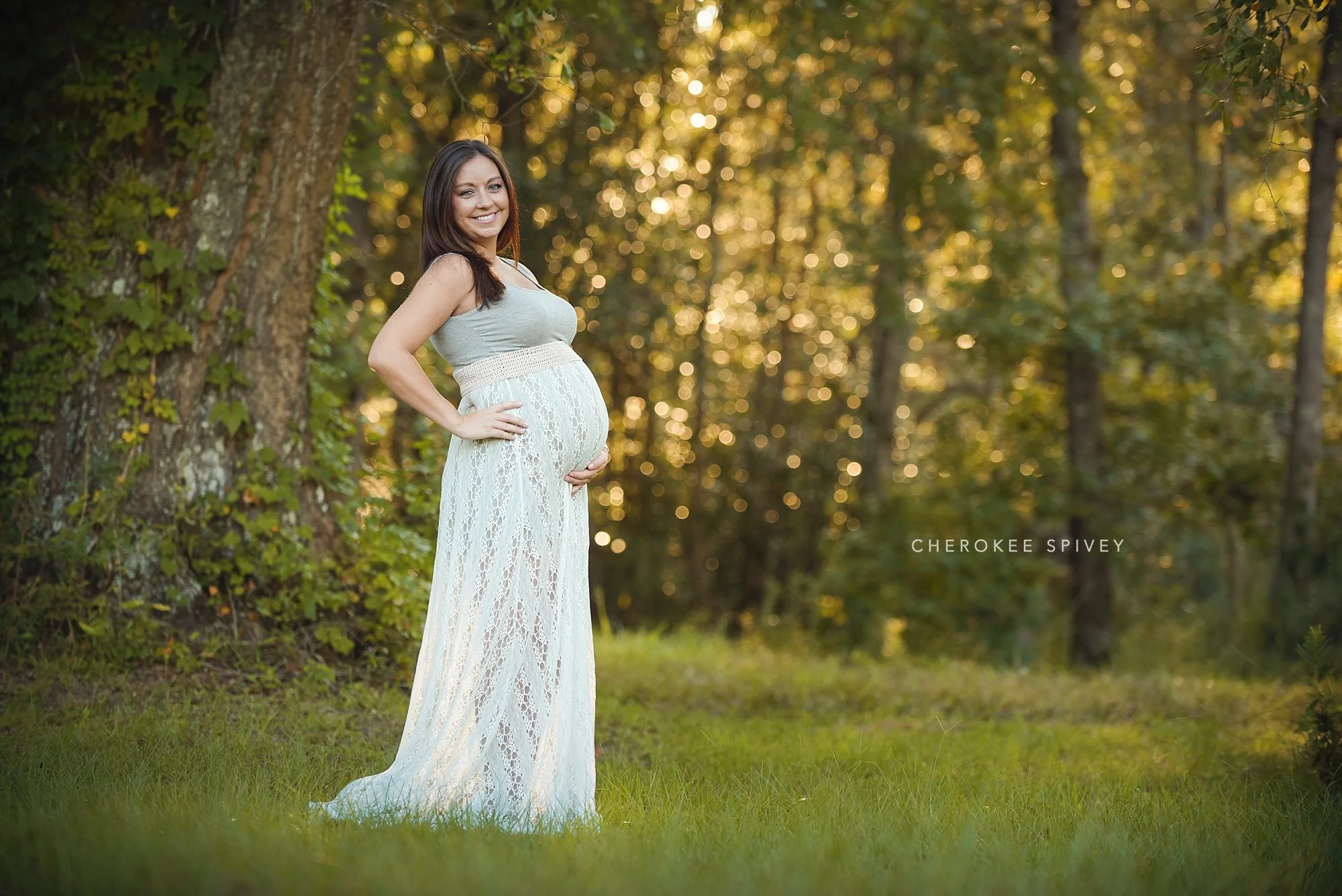 A lovely pregnant woman in a white lace gown, stands near a huge oak tree in the forest, proudly holding her big, full, belly.