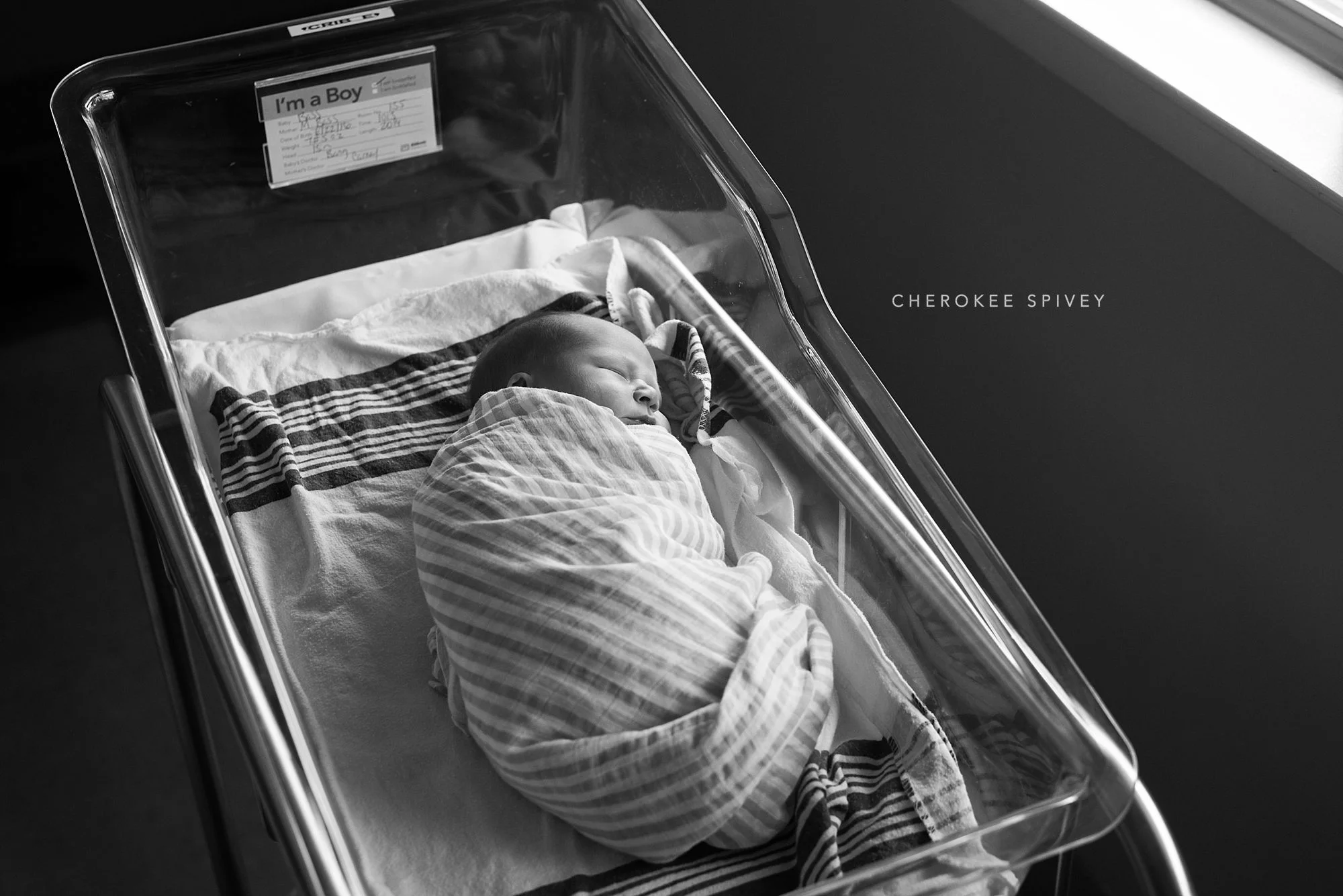 A precious newborn wrapped in a flannel blanket is laying in a clear hospital bassinet, sleeping. Above his head is a sign that says, "I"m a boy".