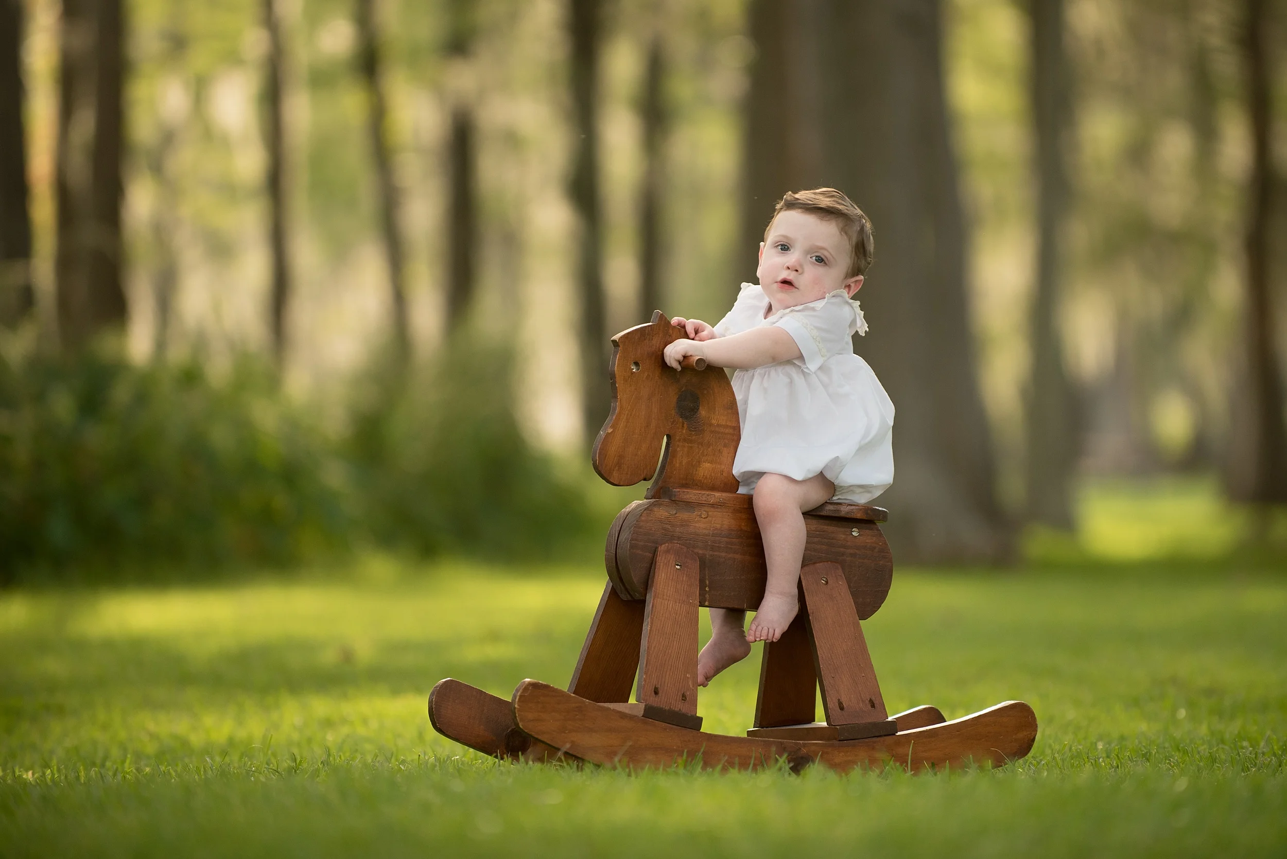 one year old infant boy in white heirloom outfit sits on an antique rocking horse outdoors