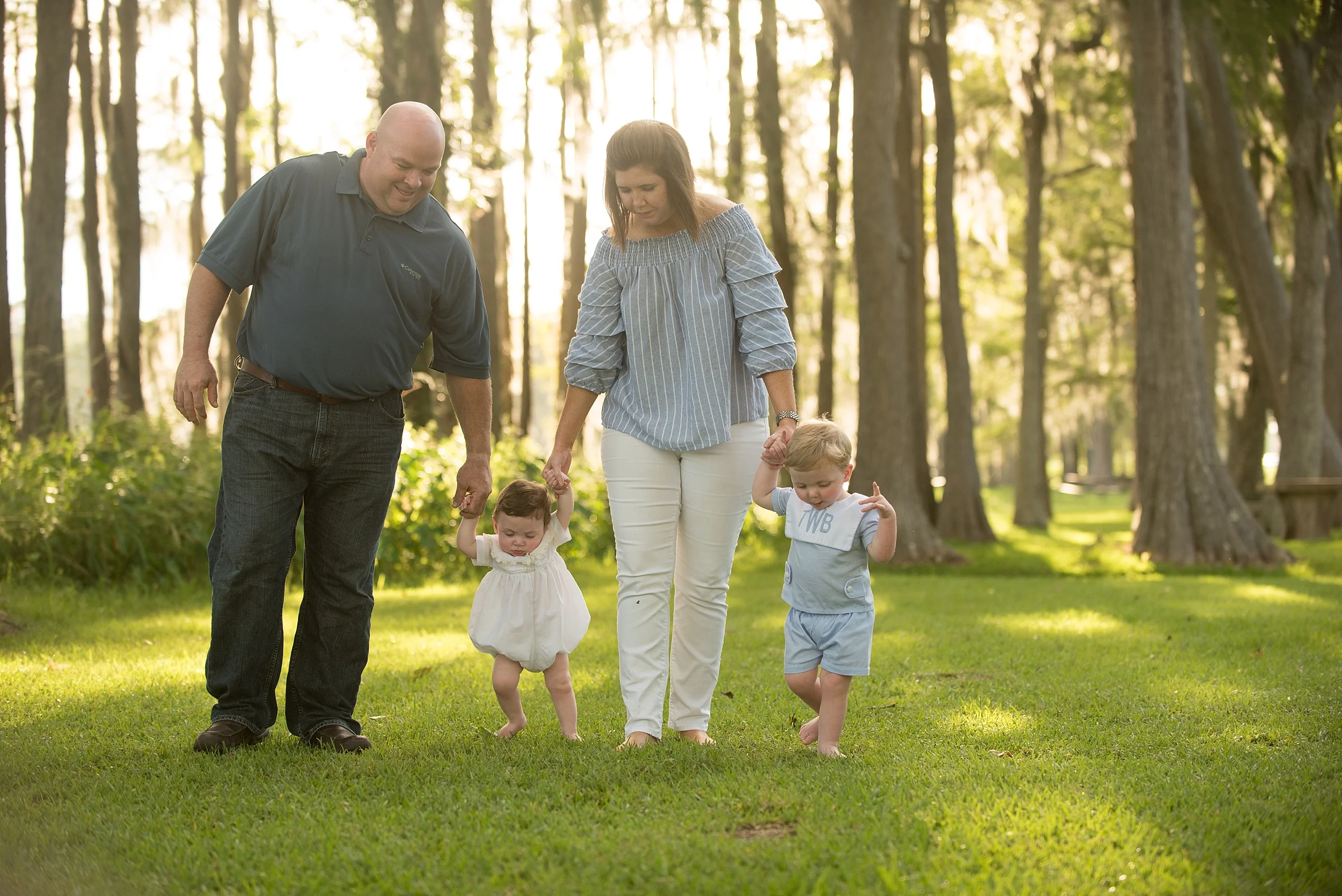 mom and dad hold hands and walk under cypress trees with their infant sons in blue heirloom outfits while golden hour sun peeks through