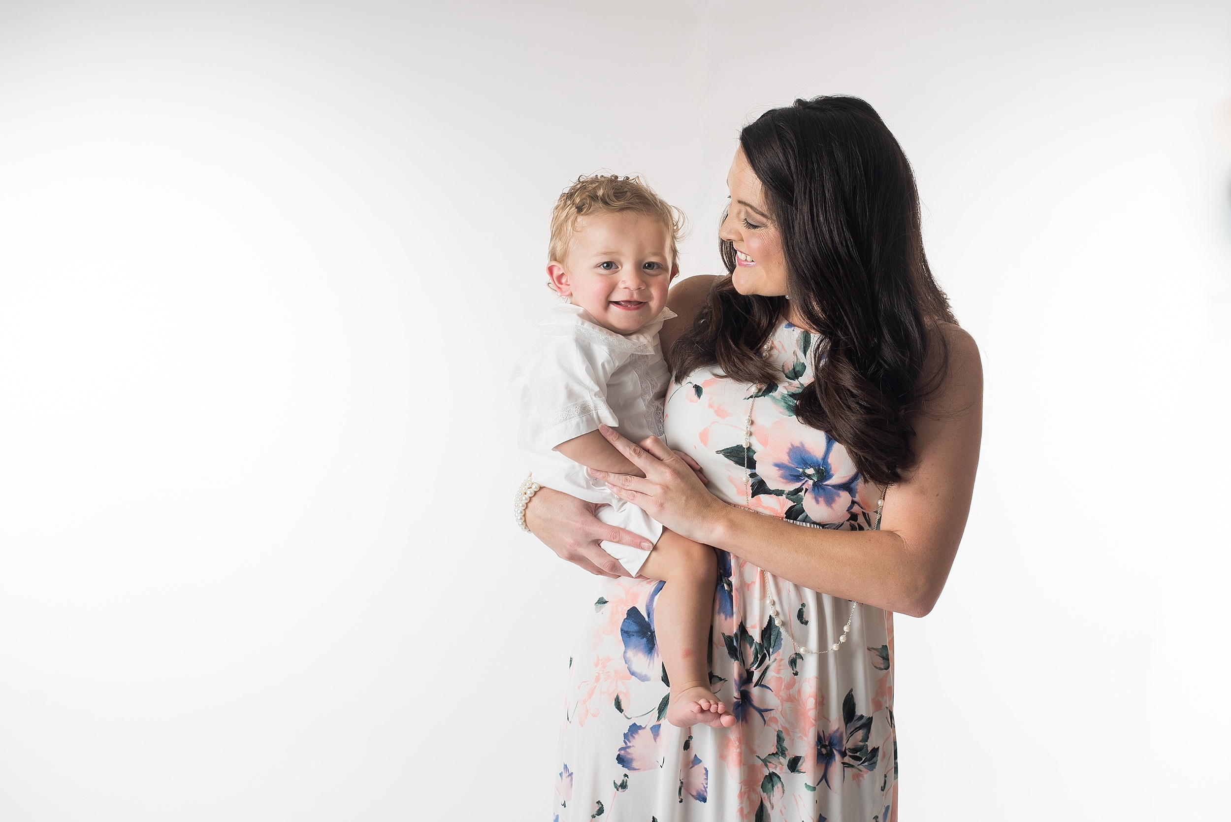 mother holds one year old boy on her hip on a white background