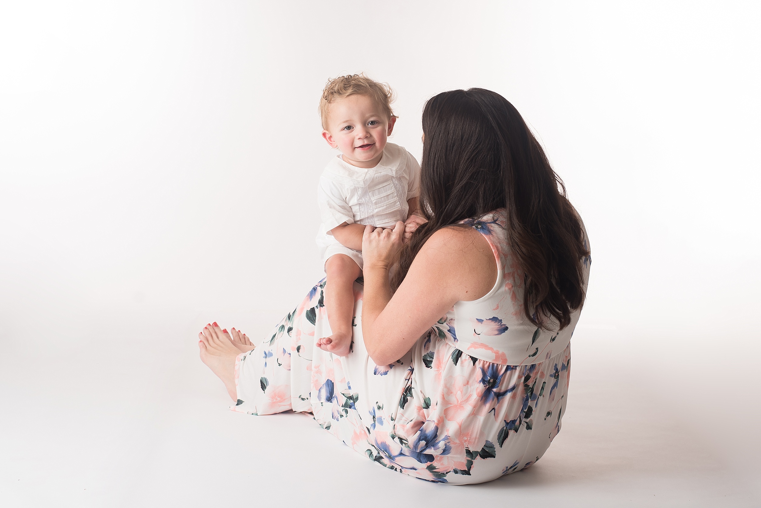 one year old boy peeks over his mom's shoulder