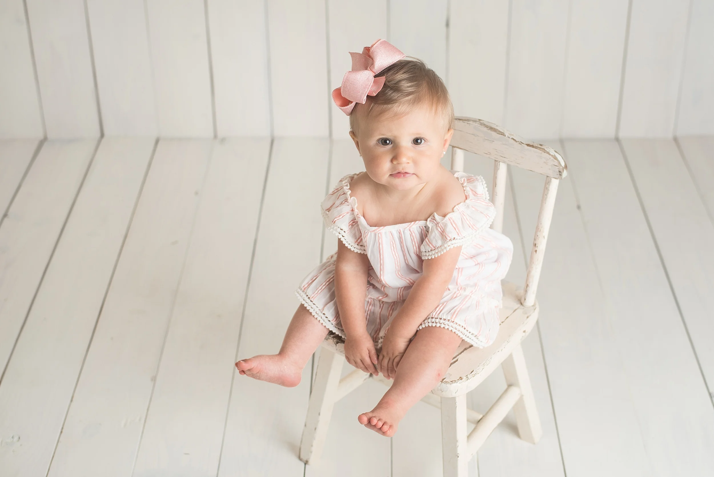 toddler girl with pink dress sits in an old white chair on a white wooden backdrop