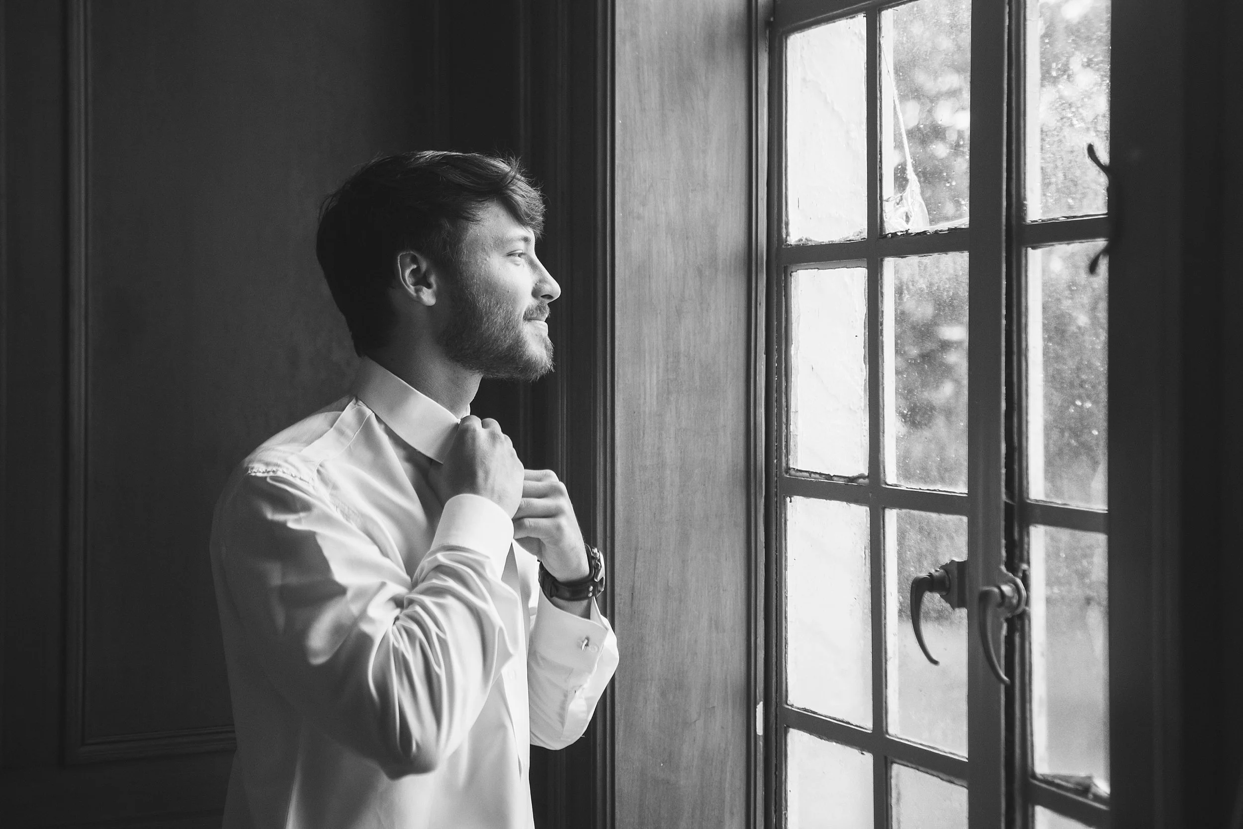 Groom fixes his tie in front of a window