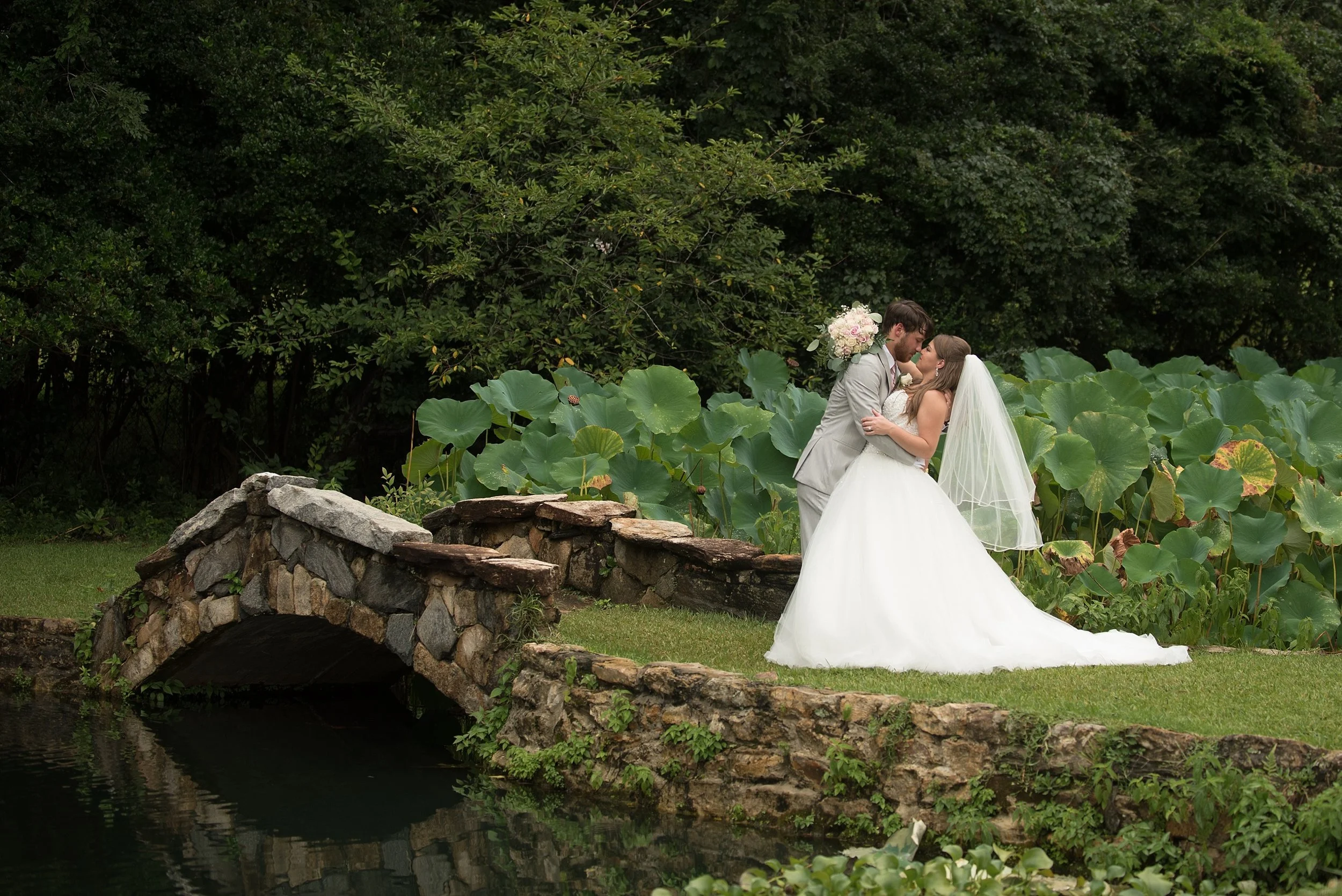 Groom dips bride and gives her a kiss beside a stone bridge