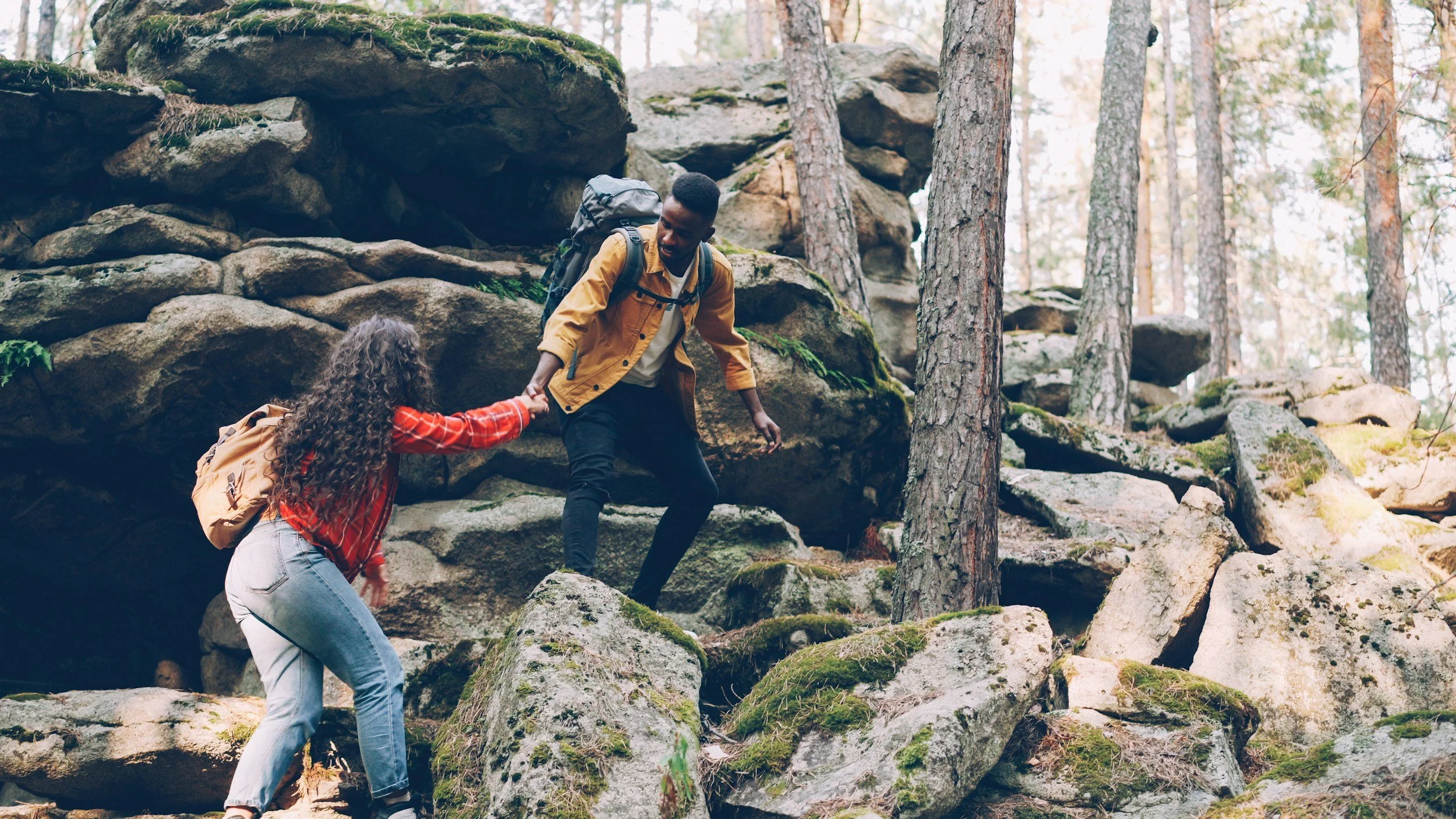 Two hikers, a man and a woman, helping each other climb over rocks in a forest.