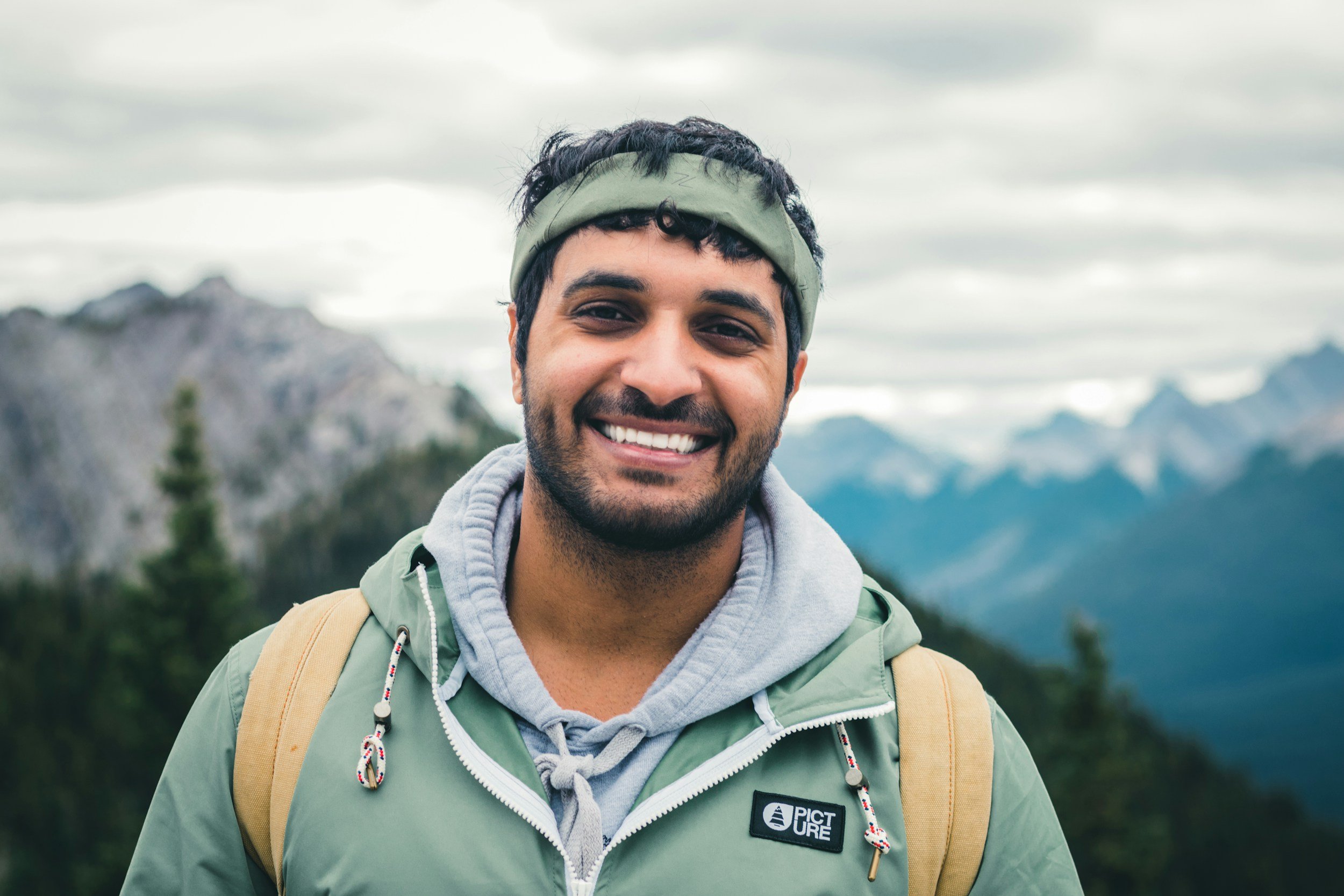 Smiling young man wearing a green jacket and gray hoodie with a backpack, standing outdoors in a mountainous area with cloudy skies.