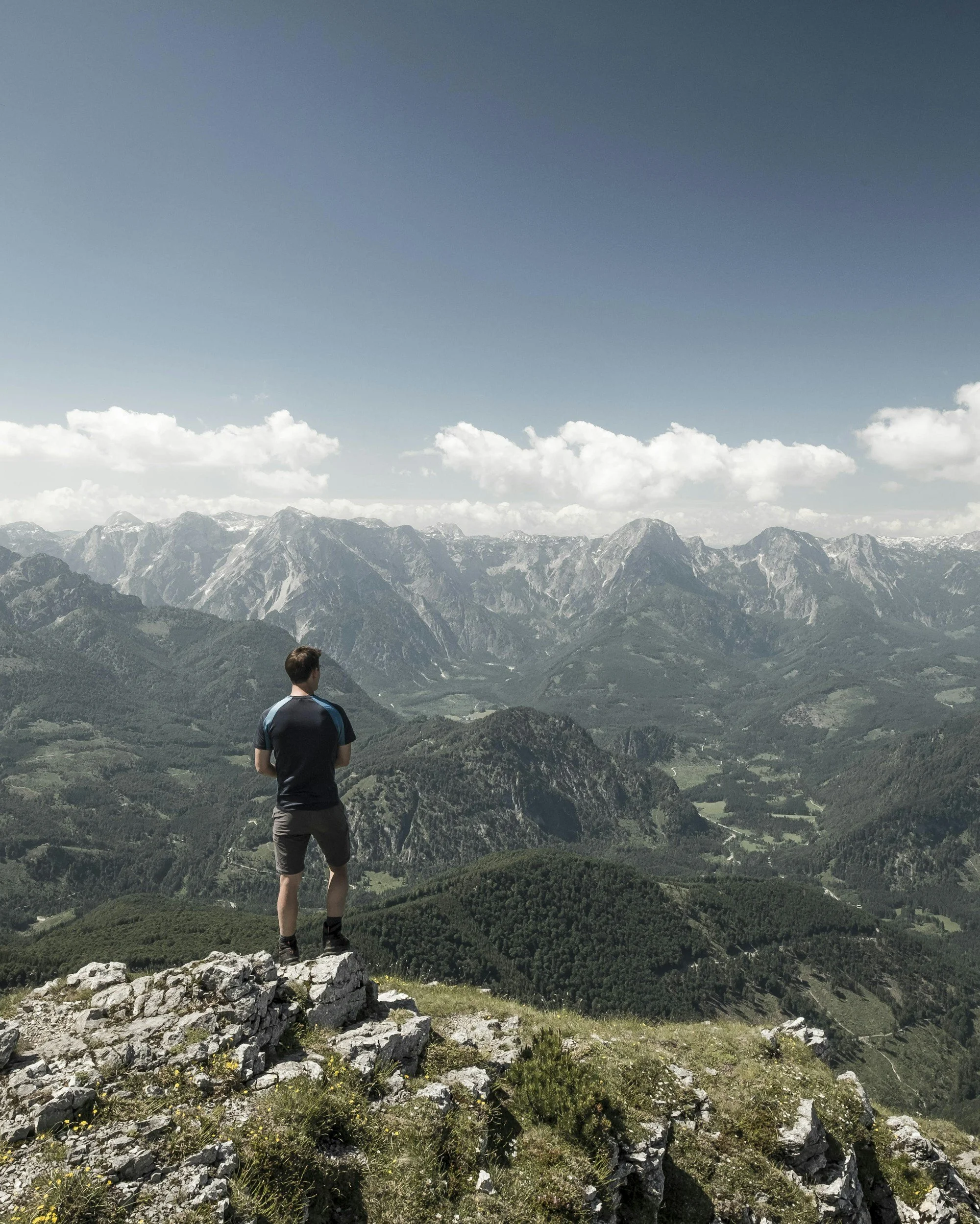 A person stands on rocky terrain overlooking a valley with mountains in the background under a partly cloudy sky.
