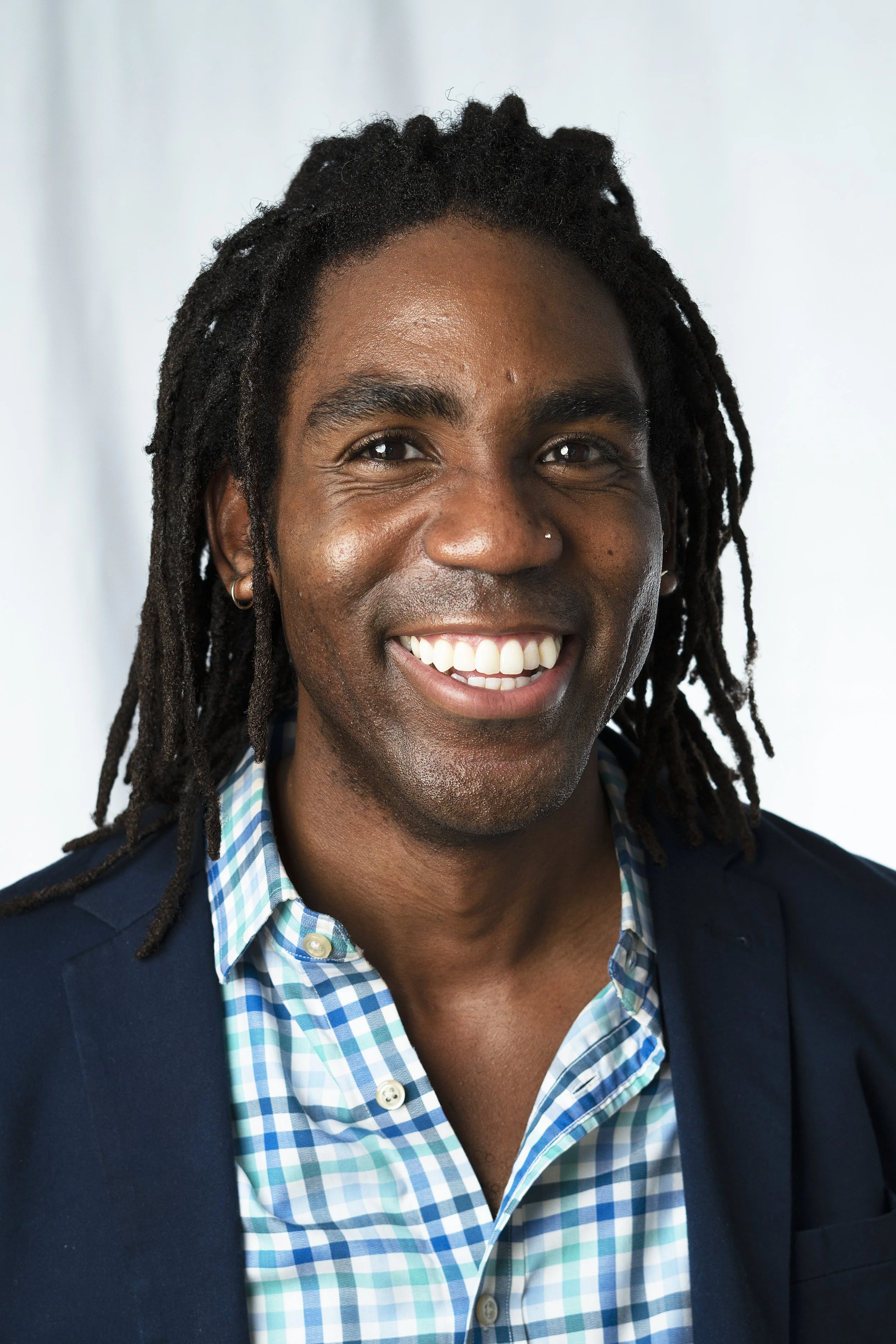 Portrait of a smiling Black man with dreadlocks, wearing a checkered blue and white shirt, a dark blazer, and silver hoop earrings, against a light background.