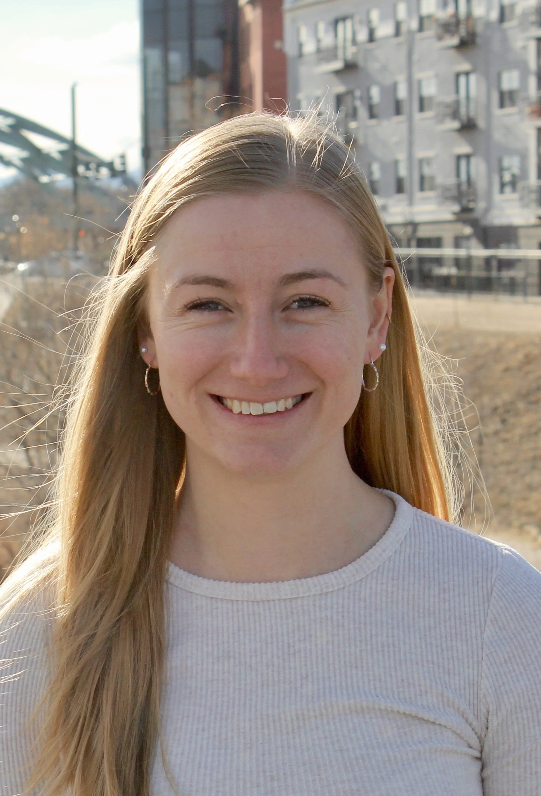 A young woman with long blonde hair smiling outdoors in front of city buildings, wearing a white ribbed shirt and hoop earrings.