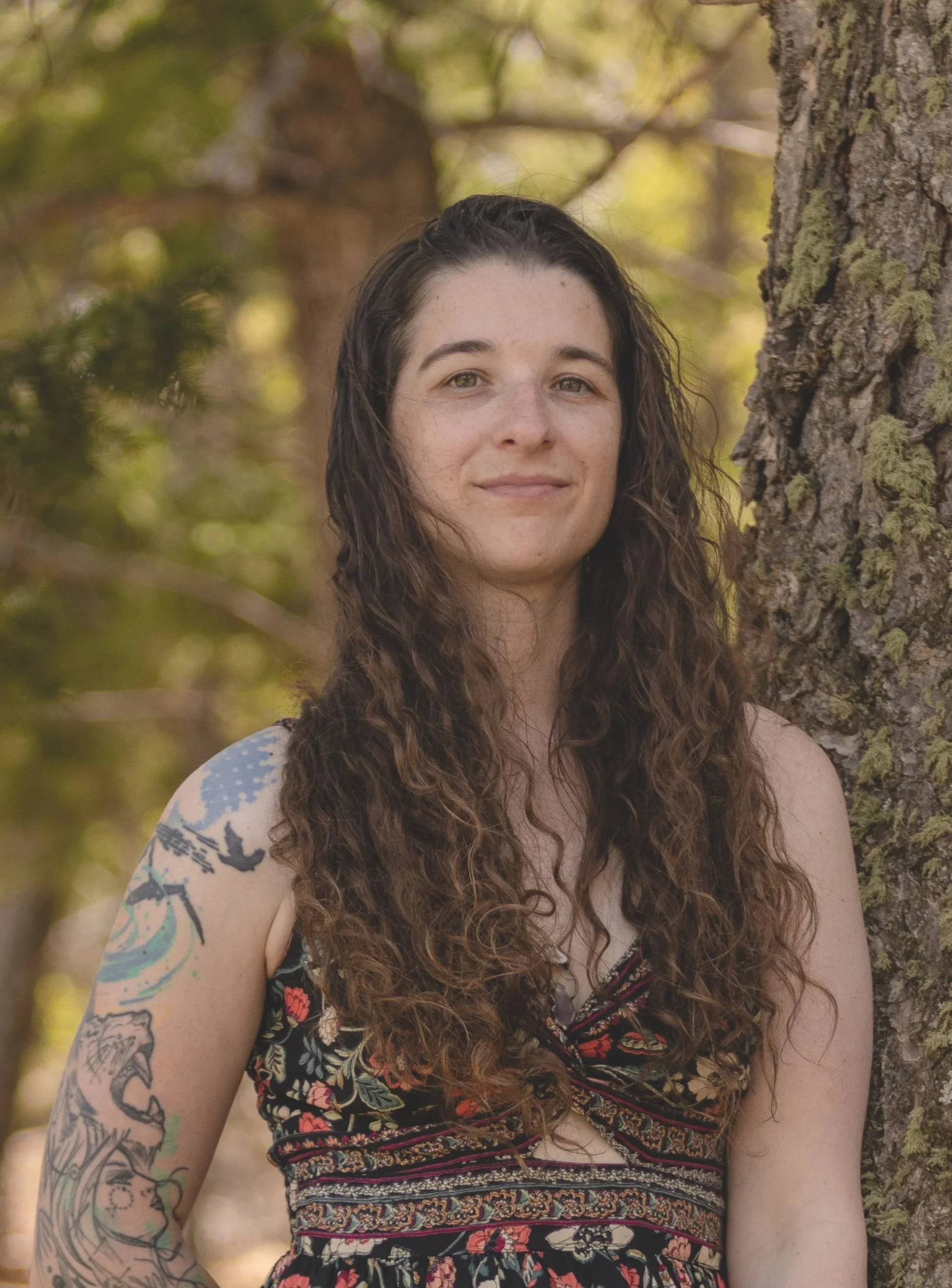 A woman with long, curly hair standing outdoors next to a tree with green leaves, wearing a patterned dress. She has tattoos on her arm and is smiling softly.