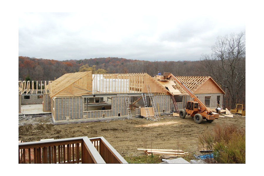 Under construction house with wooden framing, construction equipment, and ladders on a dirt lot, surrounded by trees with fall foliage.