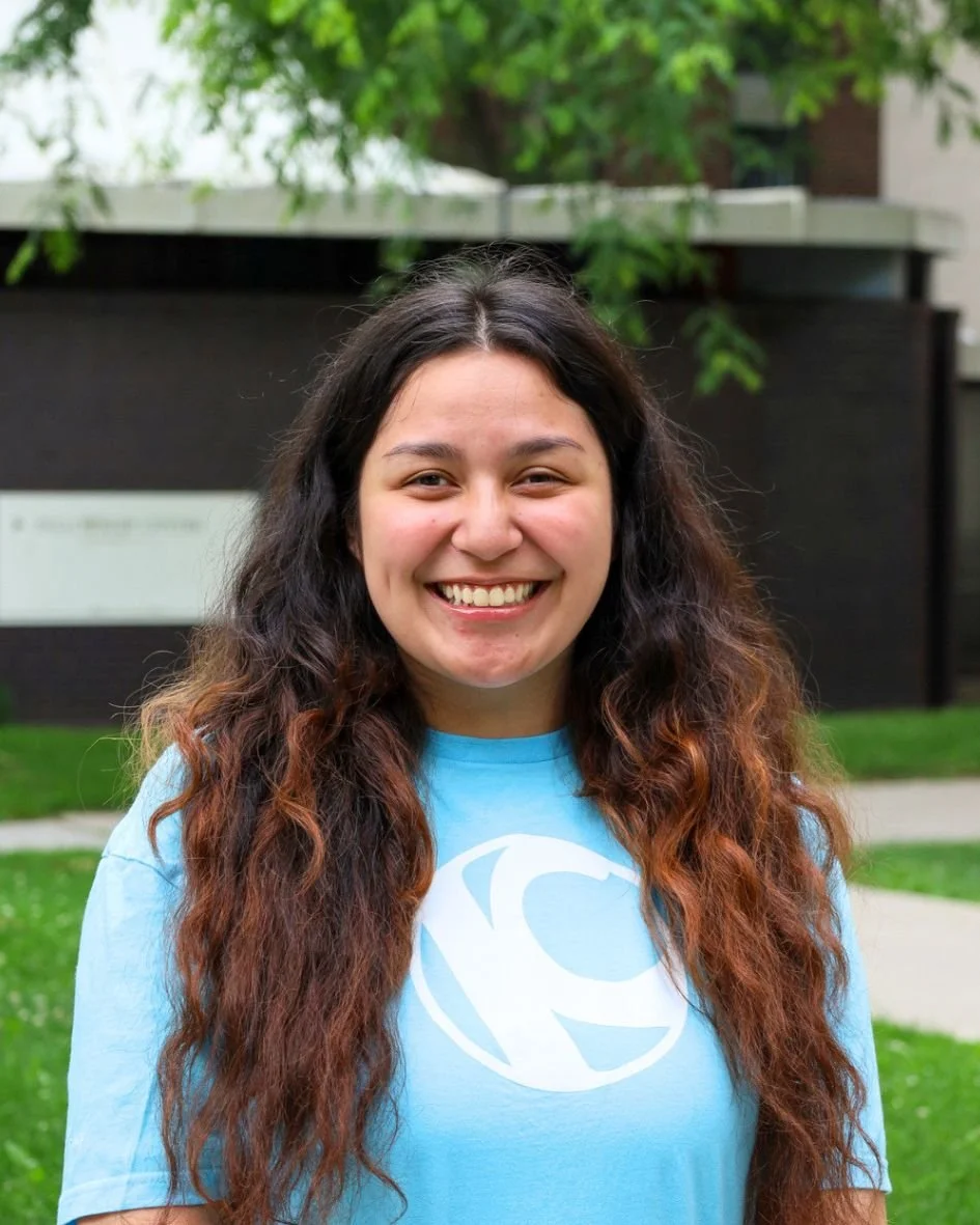 A young woman smiling outdoors, wearing a light blue shirt with a white logo, standing in front of green trees and a modern building.