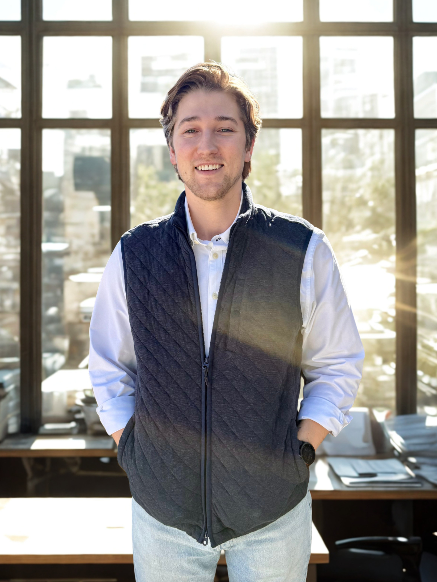 Young man with brown hair and light skin smiling, wearing a white shirt and a black quilted vest, standing in front of a large window with cityscape view.