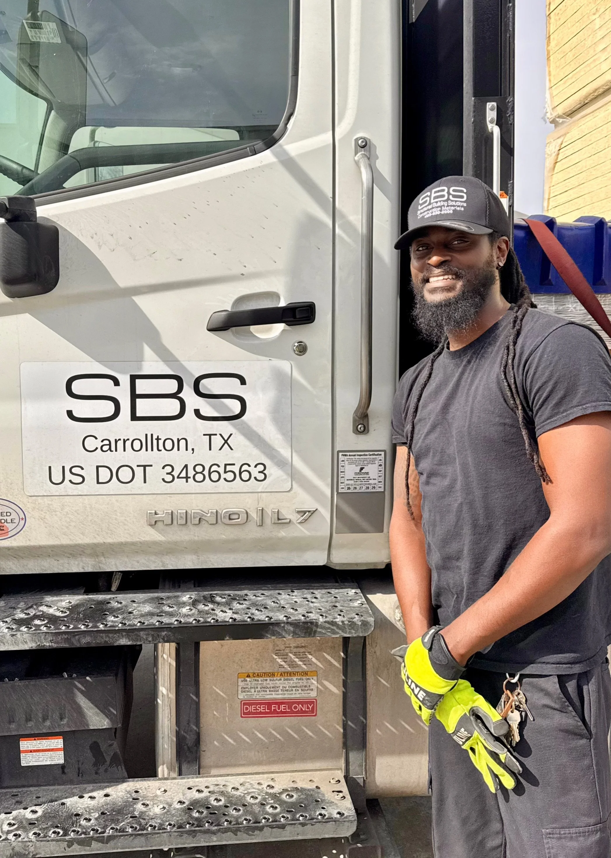 A man with a beard and long hair wearing a black cap, black T-shirt, and yellow protective gloves standing next to a white diesel fuel truck with company logo 'SBS' and location 'Carrollton, TX' painted on the side.